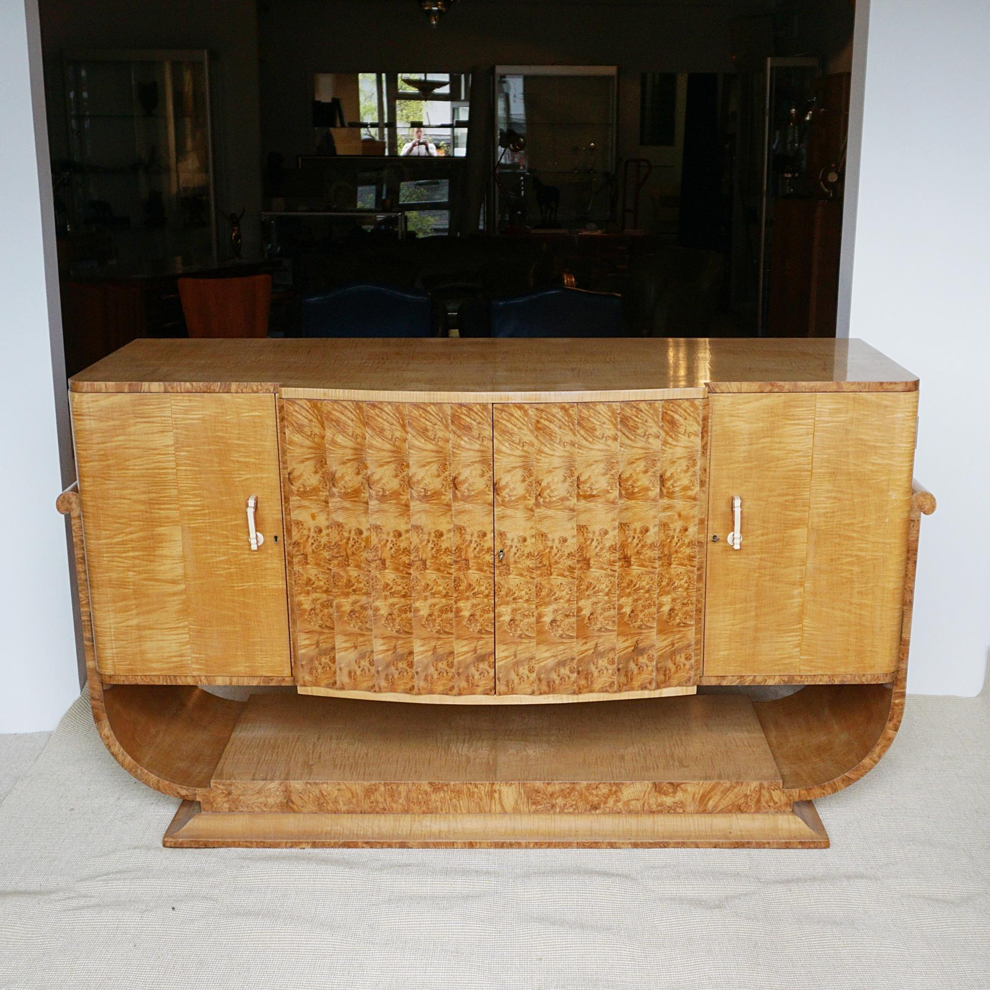 An Art Deco Sideboard by Harry & Lou Epstein. Burr walnut and satin birch veneered on solid mahogany. Central cupboard opens to reveal four mahogany lined shelves. Flanked by a single shelved cupboard to the left hand side and a shelved cupboard to