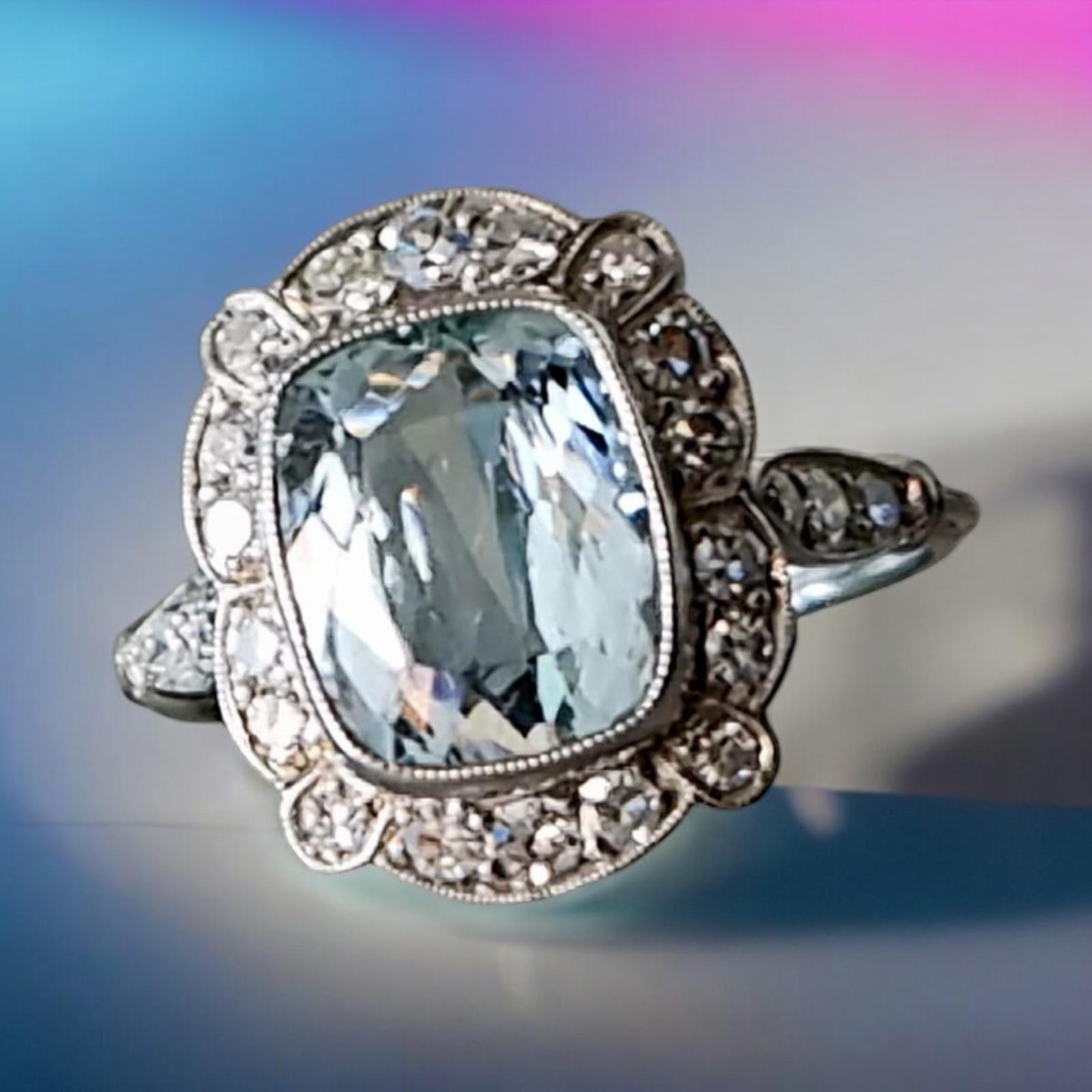 Original Art Deco Natural Aquamarine and Diamond Cluster Ring mounted in Platinum.
A refreshingly cool light blue hue colors the billowy cushion-cut aquamarine that glistens. The gemstone, resting on a double beveled bezel with milgrain edges, is