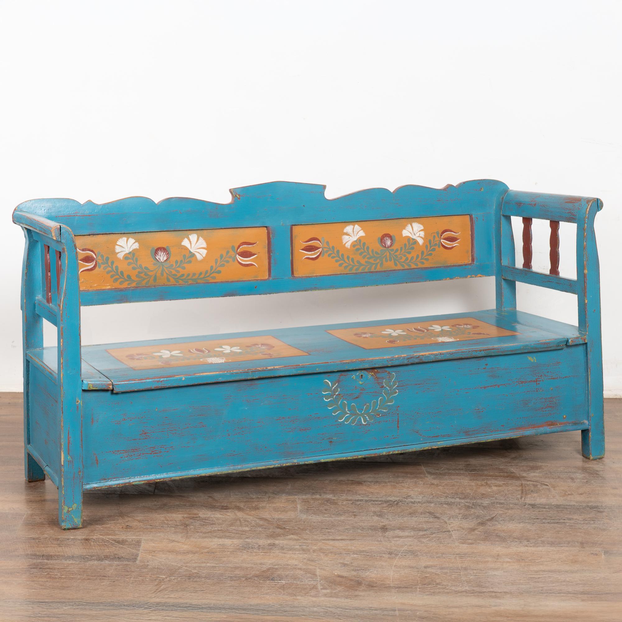 Original hand painted folk art pine bench with colorful blue background and floral motif in painted panels.
Solid back, slightly curved armrests and hinged bench seat revealing interior storage. Note on interior of bench painted names and date of