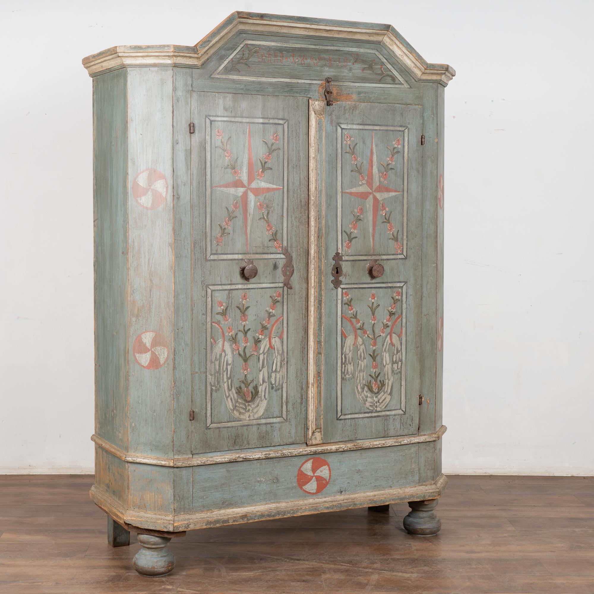 This lovely pine armoire still maintains the quality and details of the original hand-painted finish with background tones of soft blue.
Note in the crown/bonnet the monogram and date of 1810.
Large stars and floral details embellish the two doors