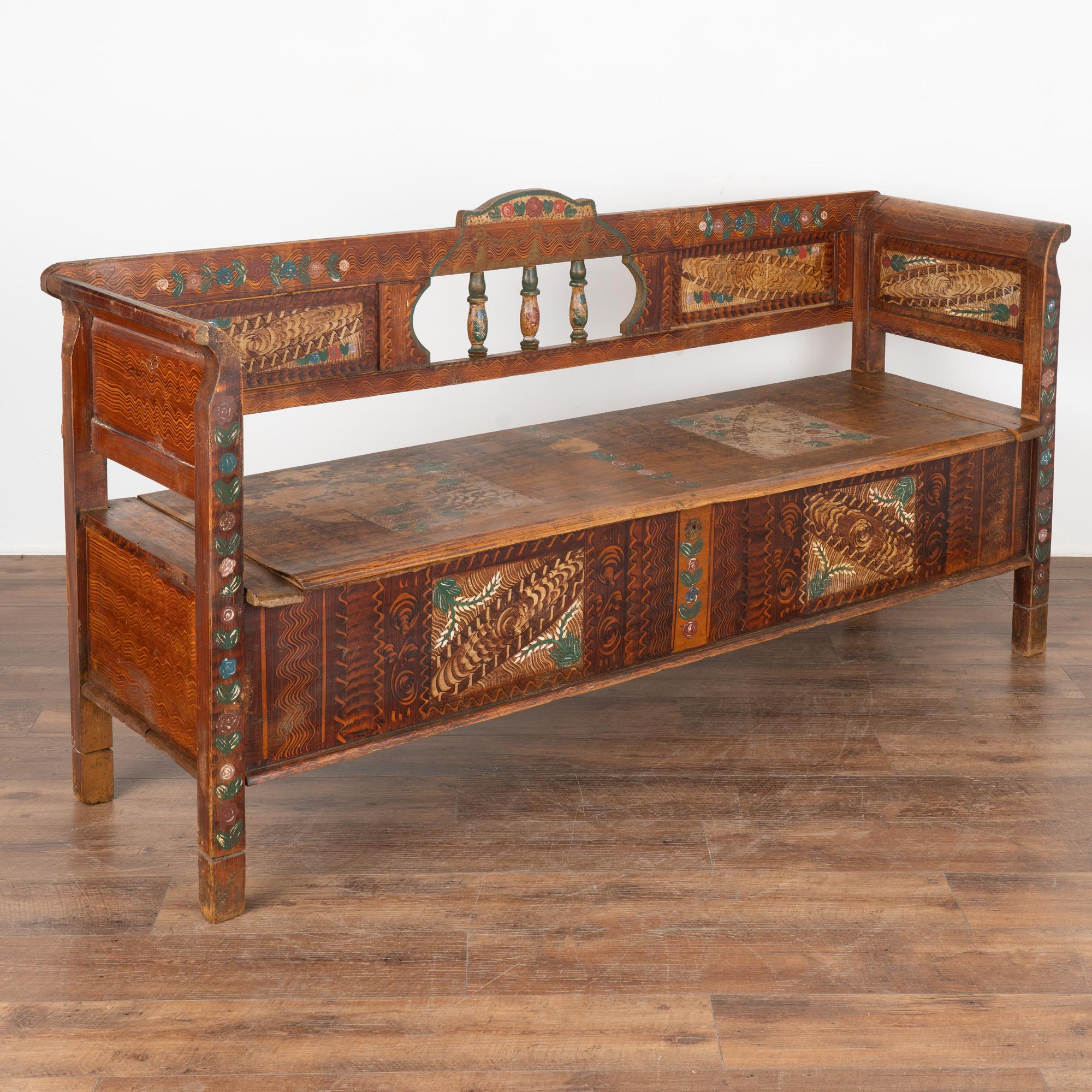 Original hand painted folk art pine bench displays traditional brown faux wood background and colorful floral motif.
Back has 3 decorative spindles, slightly curved armrests and hinged bench seat revealing interior storage. 
Restored, cleaned and