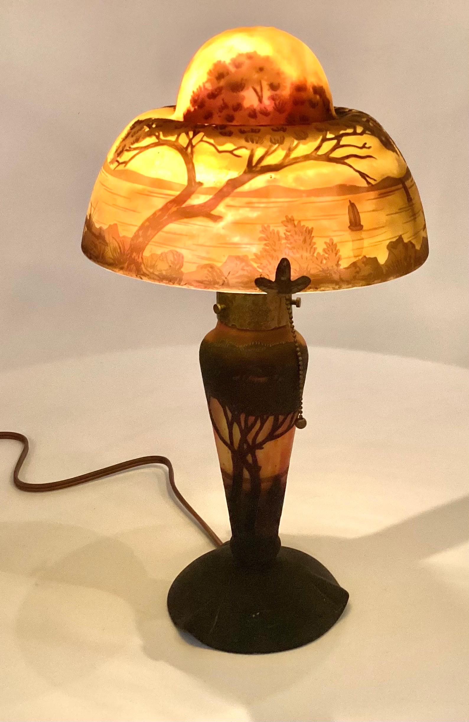 Une lampe de table originale de Daum Nancy en verre camée coloré. La superposition de couleurs sombres contraste avec le fond orange sur or. L'ombre est une scène de lac tranquille avec deux grands arbres et 5 voiliers, la base représentant des