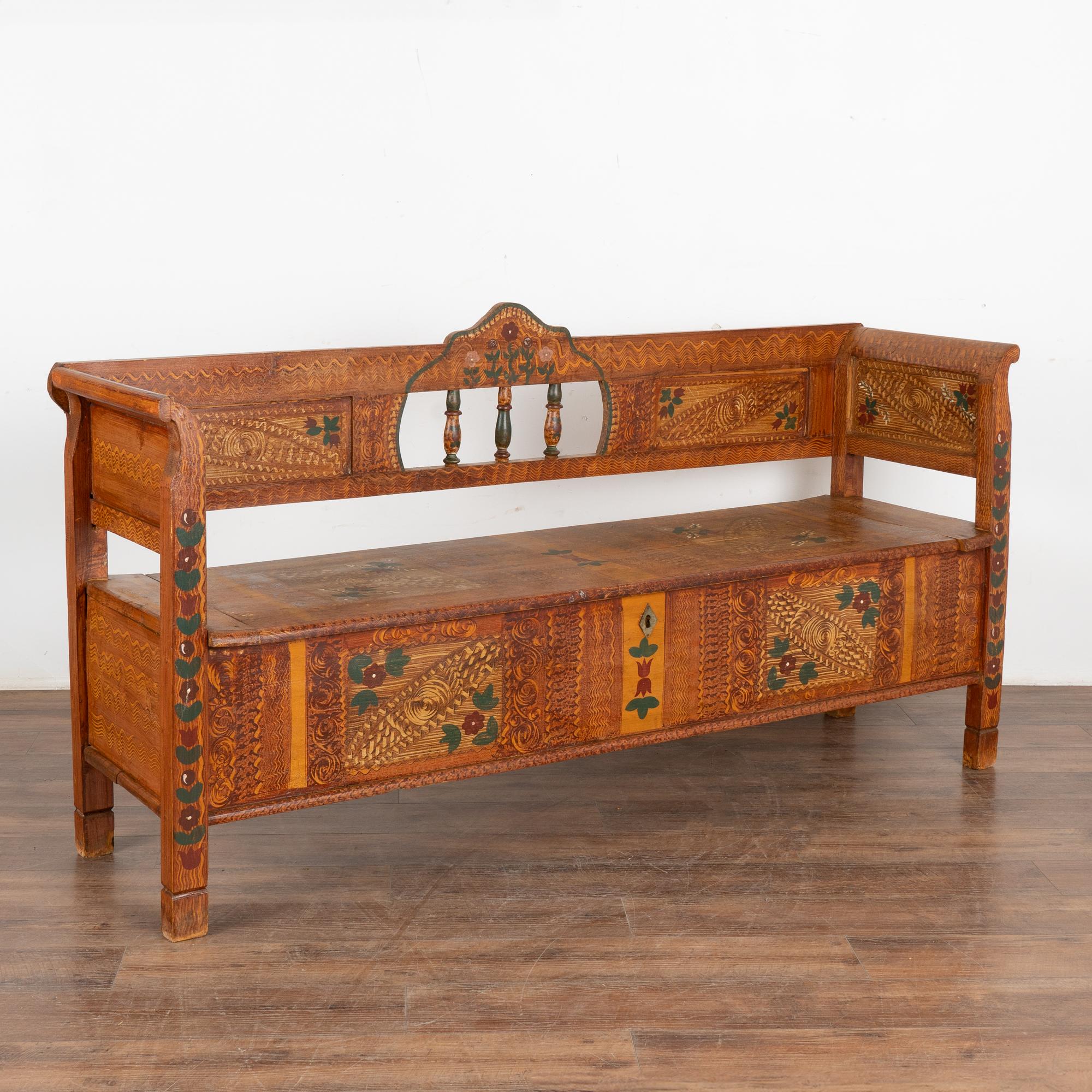 El original banco de pino de arte popular pintado a mano muestra un fondo tradicional de imitación de madera marrón y un colorido motivo floral.
El respaldo tiene 3 husillos decorativos, reposabrazos ligeramente curvados y asiento abatible que deja