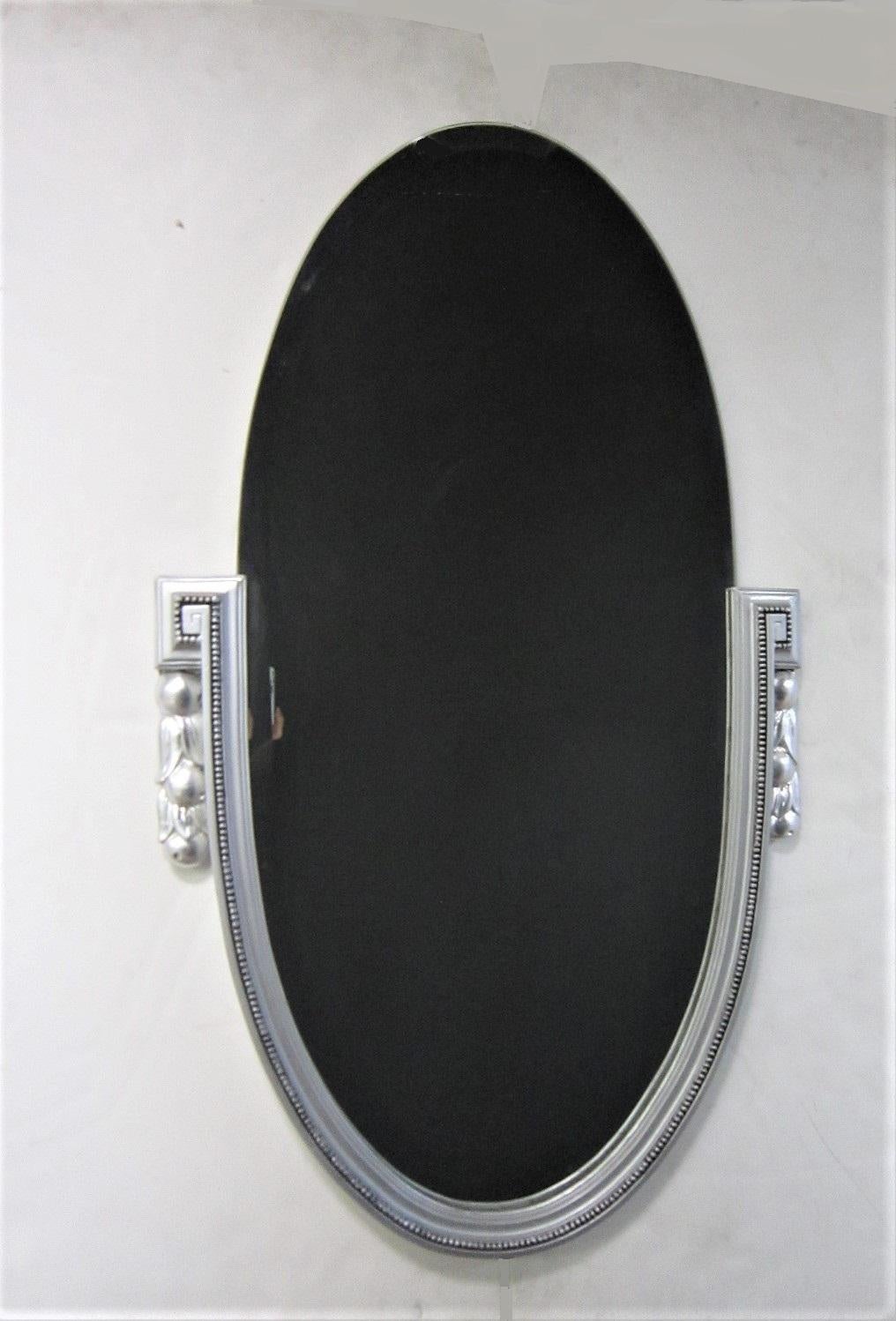 An elegant silvered wood and gesso French Art Deco elongated oval mirror with beveled mirror inset. The sides flanking either end of the widely beveled mirror feature a Greek key design with a naturalistic motif of flowers or berries and leaves