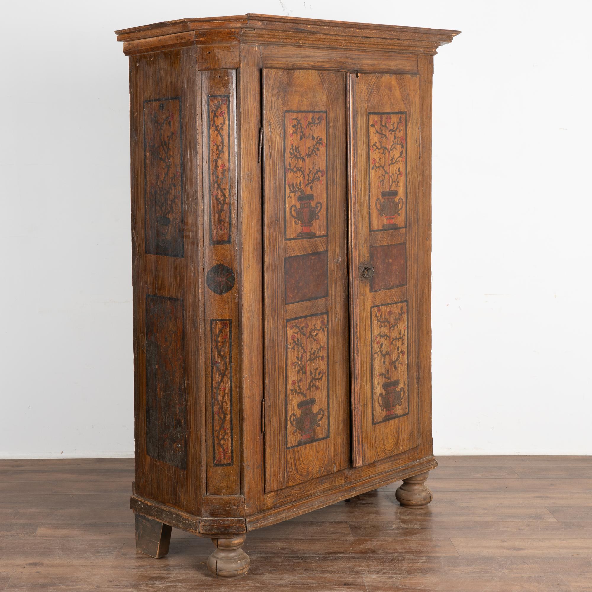 Cette armoire en pin peint conserve la qualité et les détails de la finition originale peinte à la main dans une palette de tons de terre.
Les motifs floraux sur le fond peint en faux bois s'inscrivent dans le style traditionnel de l'art populaire