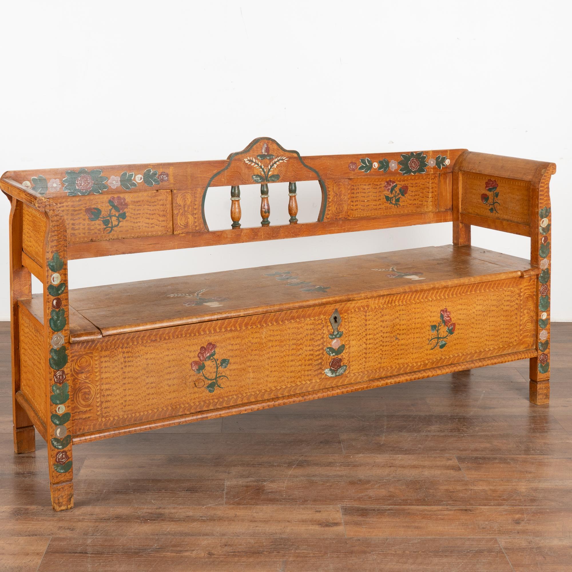 Ce banc en pin, peint à la main, présente un fond traditionnel en faux bois marron et un motif floral coloré.
Le dossier comporte 3 fuseaux décoratifs, des accoudoirs légèrement incurvés et une banquette articulée révélant un espace de rangement