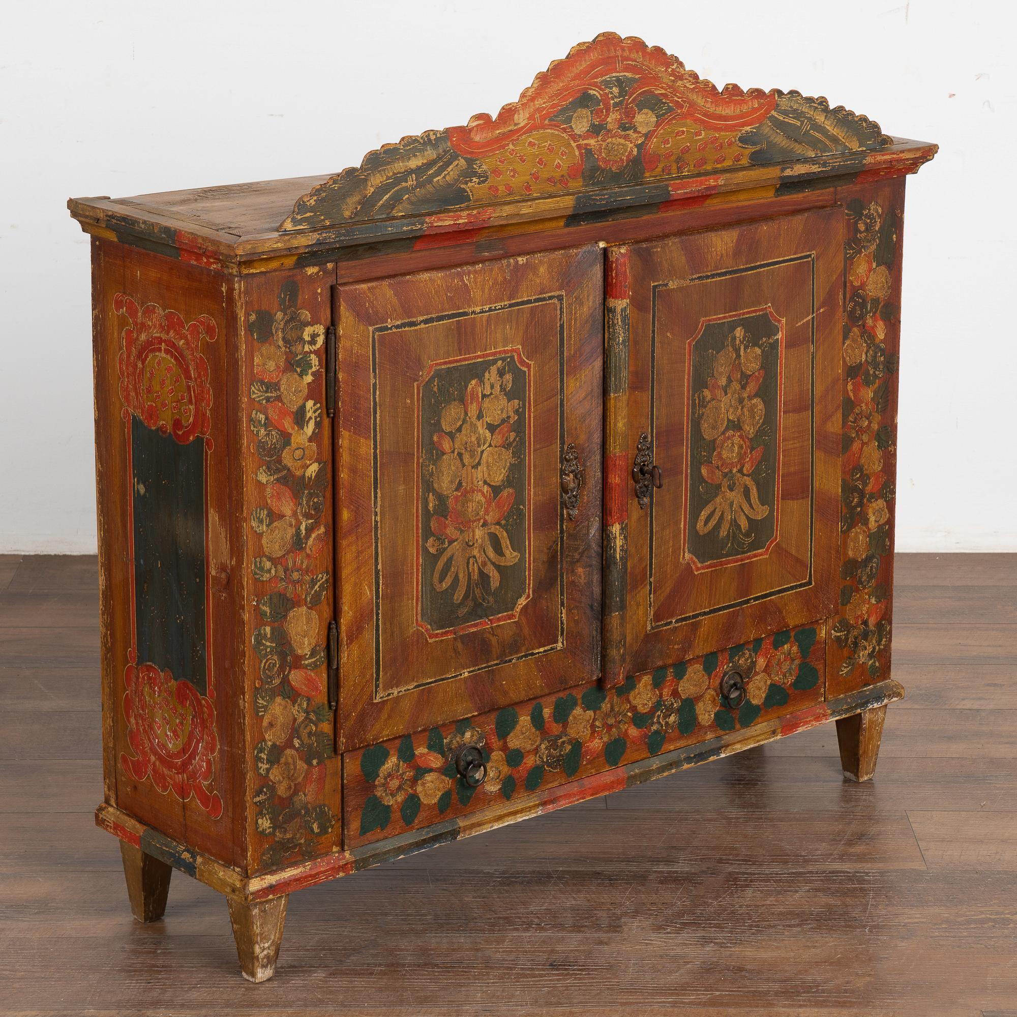 The colorful painted finish is all original on this delightful small cabinet from Austria resting on tapered feet. 
Tones of orange, red, brown, yellow, ocher and green are all blended in the hand-painted florals and flourishes that are seen
