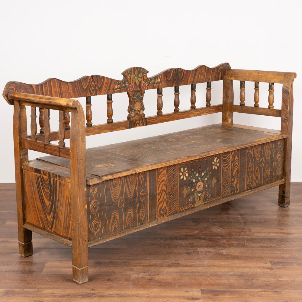 Original hand painted folk art pine bench displays traditional brown faux wood background and colorful floral motif.
Traditional spindle back, curved armrests and hinged bench seat revealing interior storage. 
Restored, cleaned and waxed, this