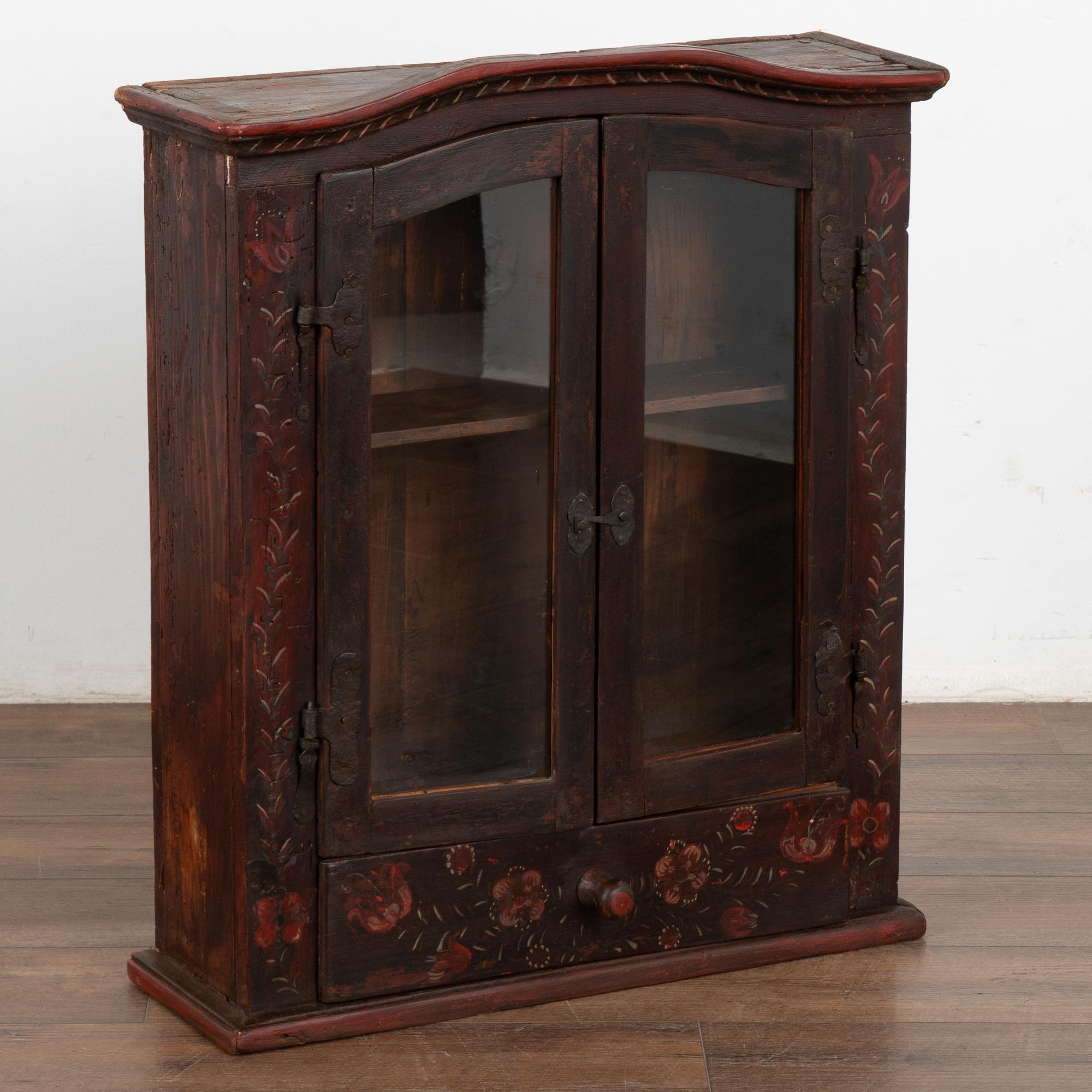 The deep patina of the antique pine is evidence of it's age in this antique hanging cabinet with original hardware. Adding to the charm is the original folk art paint that can still be seen outlined on the door panel and lower drawer. Red flowers