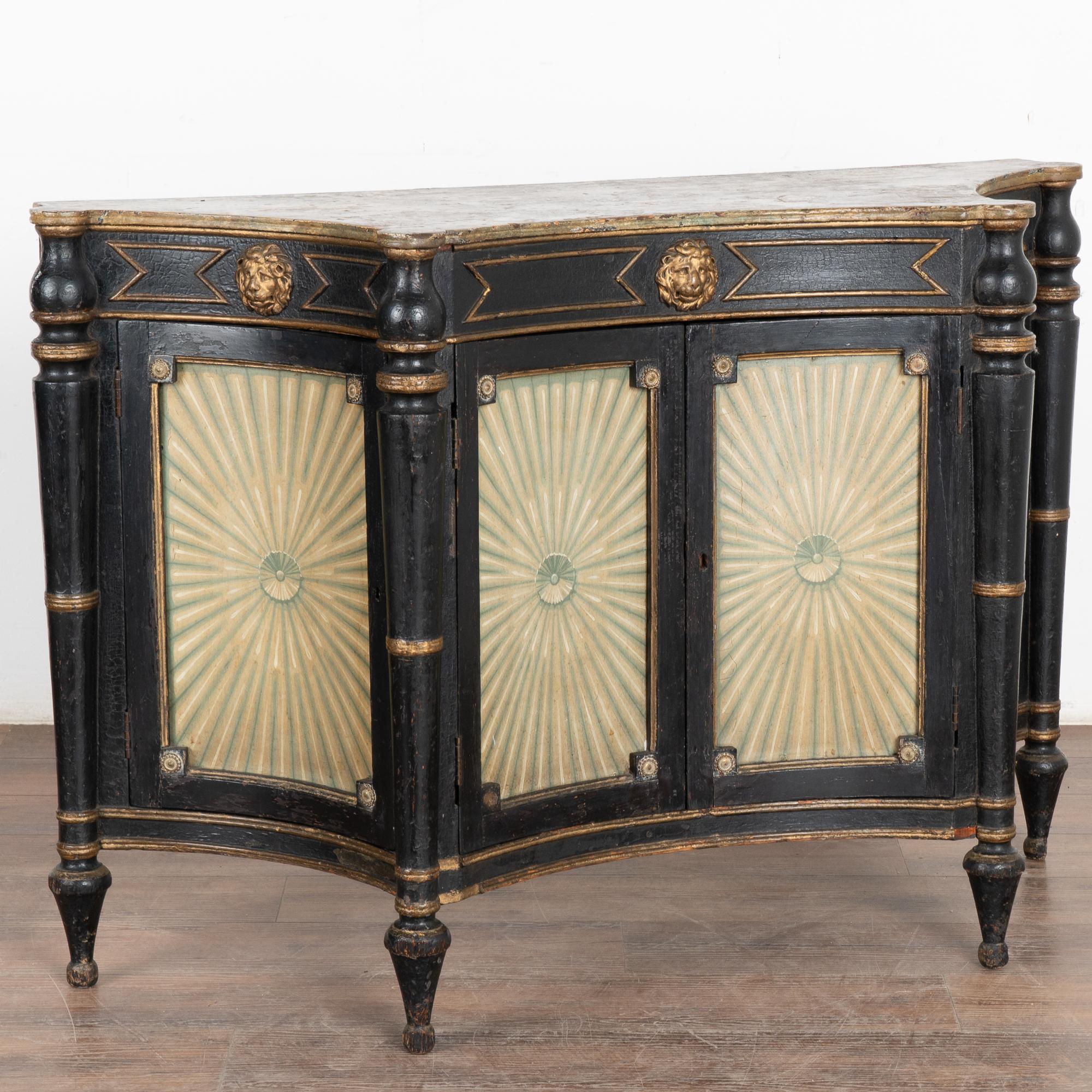 This striking late empire Italian sideboard has black painted and gilded wood, faux marble top, and four mesmerizing painted concave doors.
Additional embellishments include the lion head faces with turned columns and feet on this 4' wide