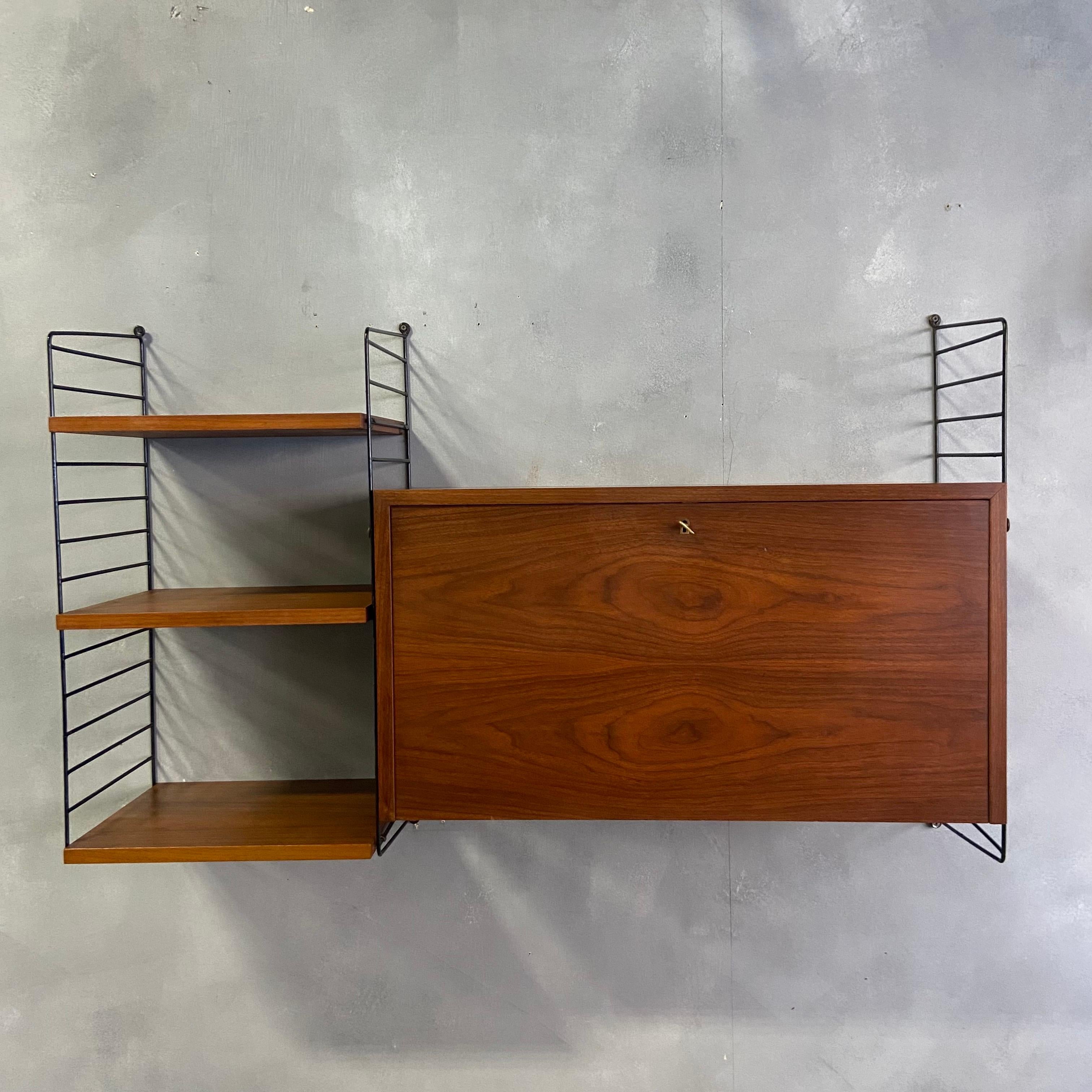 Beautiful Patina on this dark teak String Unit original from 1950. Featuring three 16'' wide shelves and a drop down cabinet / secretary concealing three dovetailed drawers in birch wood. All parts are adjustable along the ladder work using the