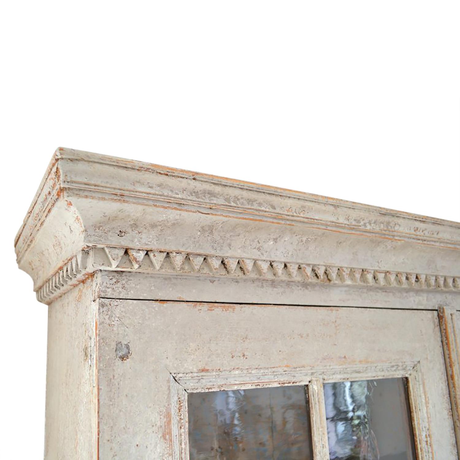 Gustavian glazed cabinet in original paint with a decorative carved pediment. This piece has two glazed doors that open to storage shelves, and two further doors opening to more storage.