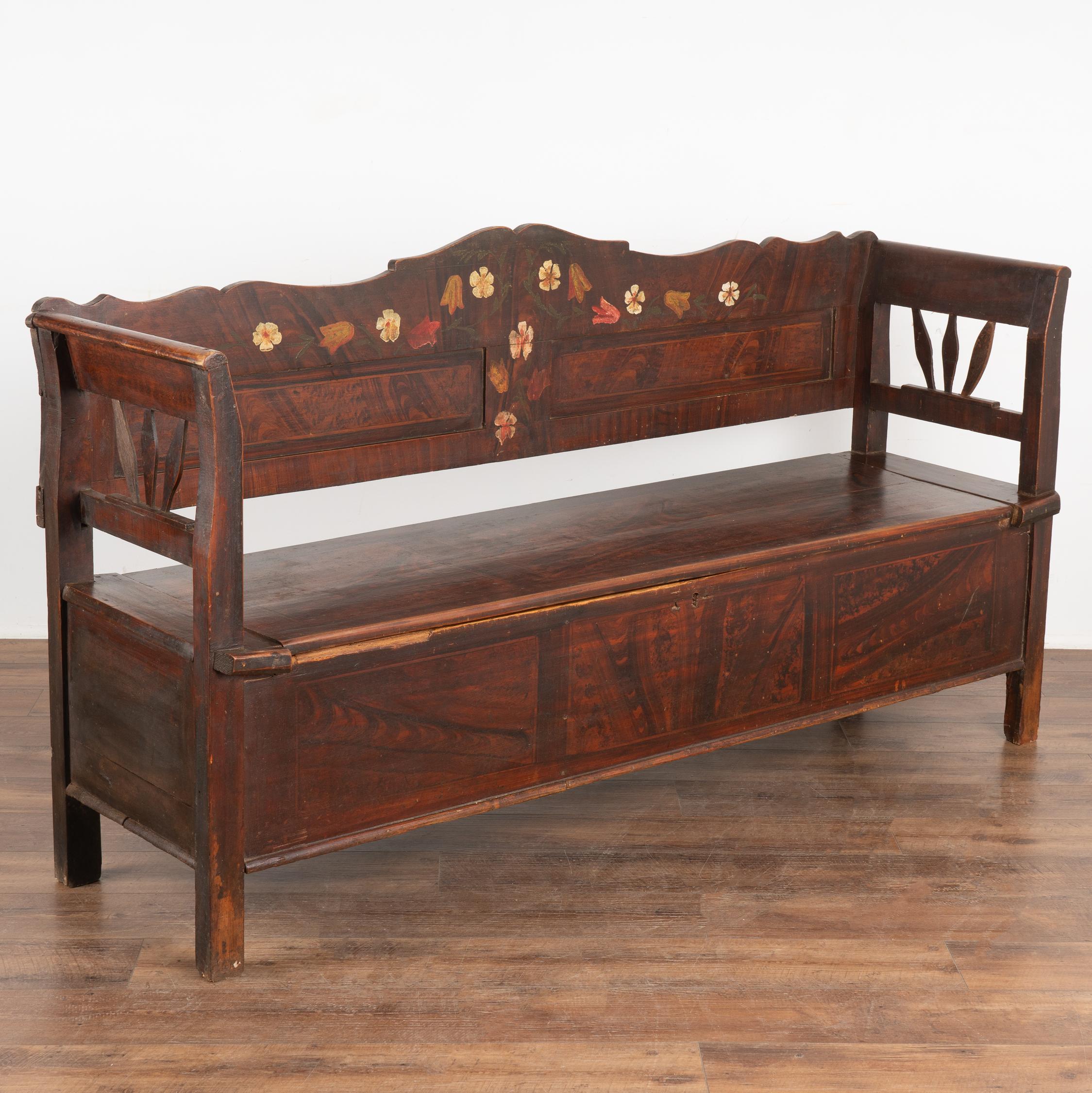 Ce banc en pin peint à la main présente un fond traditionnel en faux bois brun et un motif floral coloré le long du dossier.
La banquette à charnières s'ouvre pour révéler un espace de rangement divisé à l'intérieur (un léger rebord au coin avant