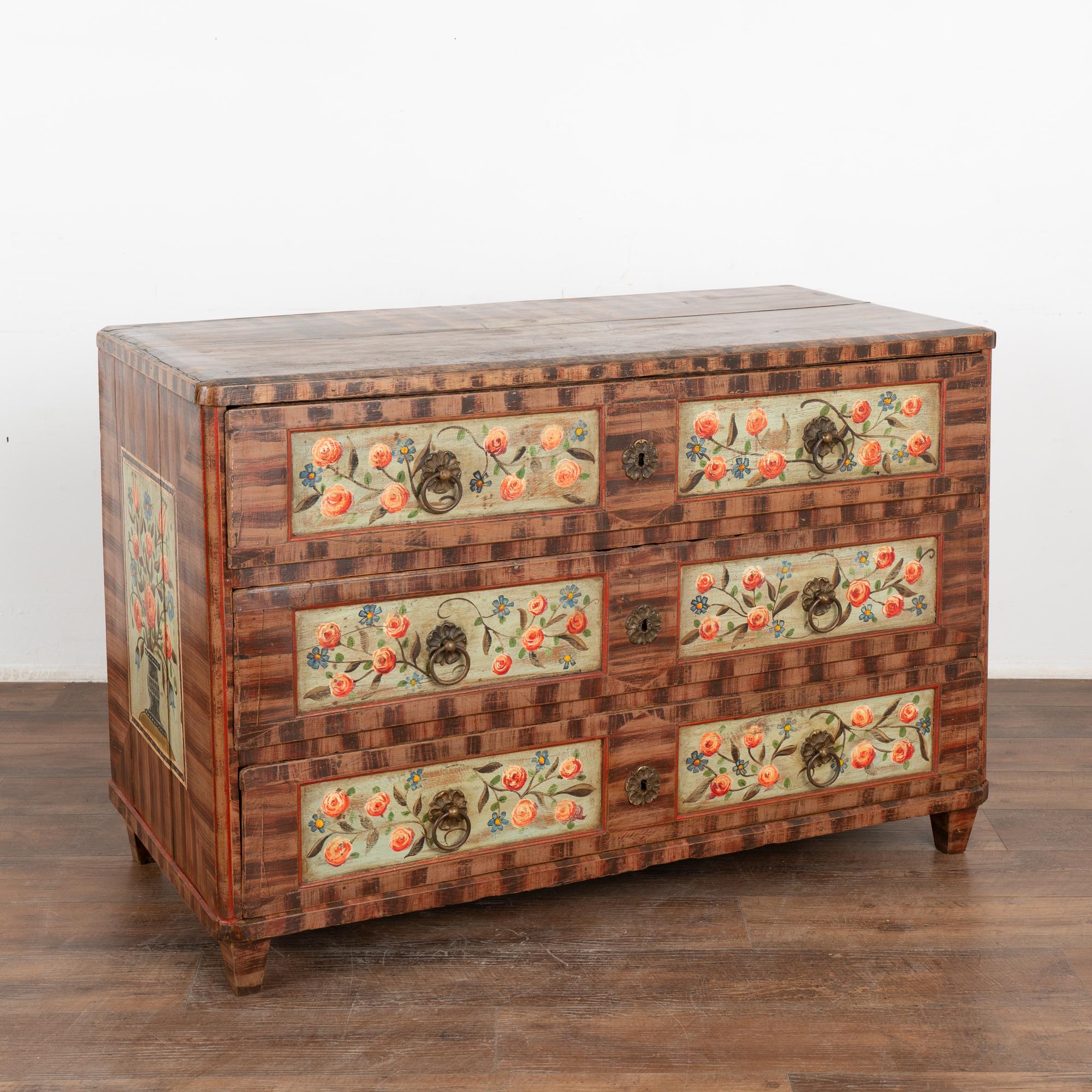 The vibrant colors are all original on this hand-painted antique chest of 3 large drawers from Hungary.
The lovely floral painted panels were a traditional folk art design of the era set against the faux wood brown finish, adding a cheerful feel to