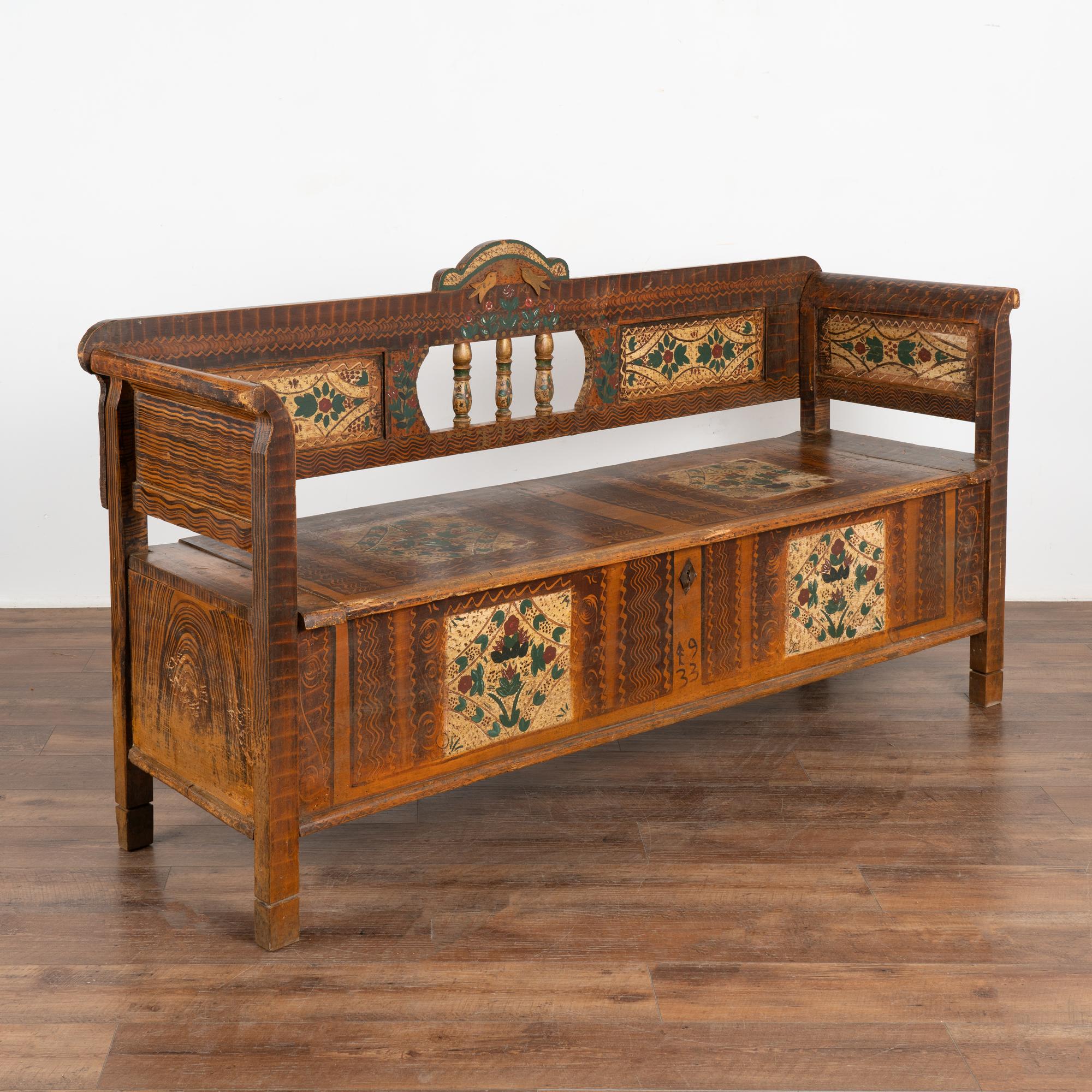 Original hand painted folk art pine bench displays exceptional traditional brown faux wood background and colorful green and red floral displays within painted panels along the front, seat, back and sides. 
Note the decorative carved birds and