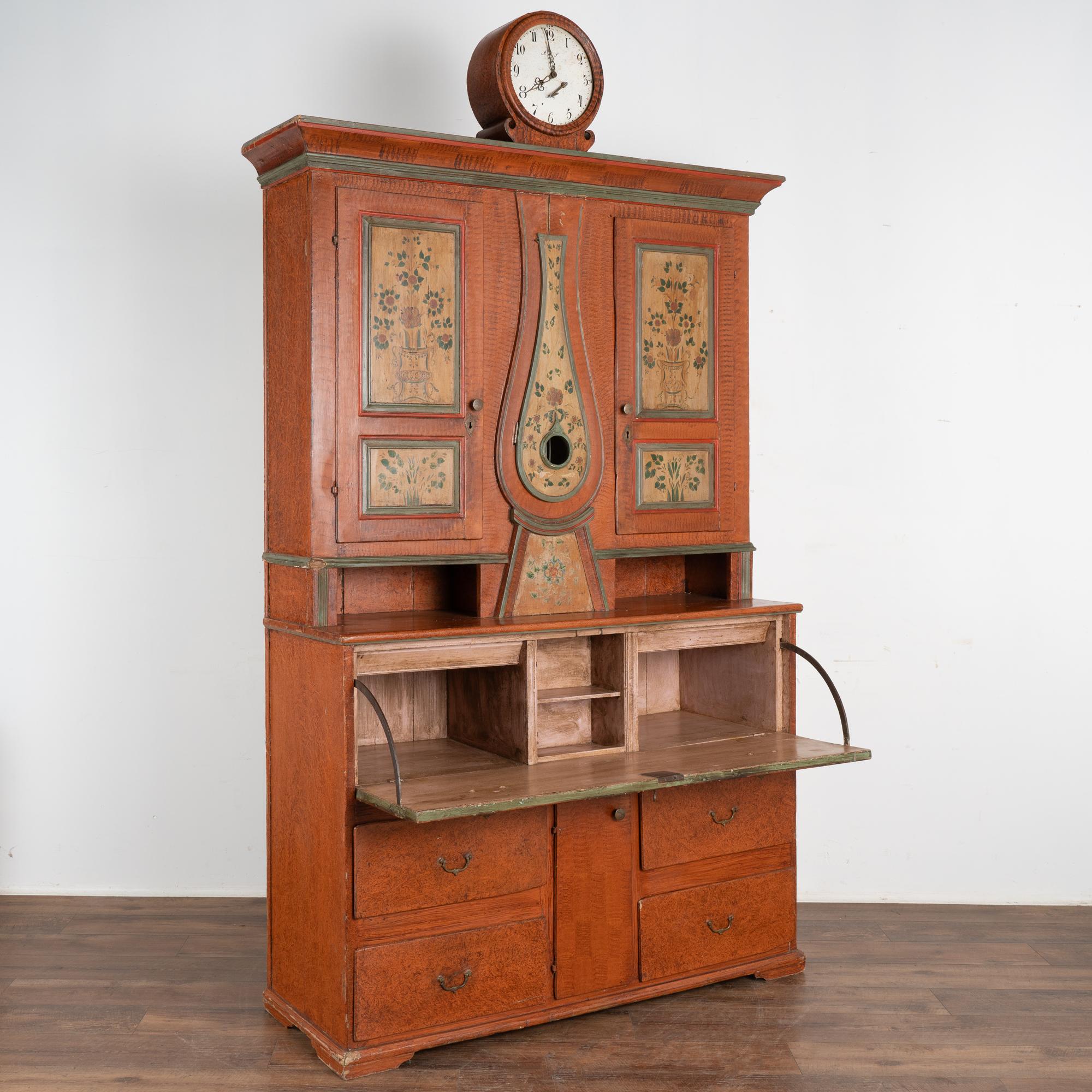 Remarkable original painted Swedish cabinet with clock and secretary with drop front desk. 
The traditional faux painted burnt umber background is balanced by the delicate hand-painted floral panels of the upper panel doors and clock body. The