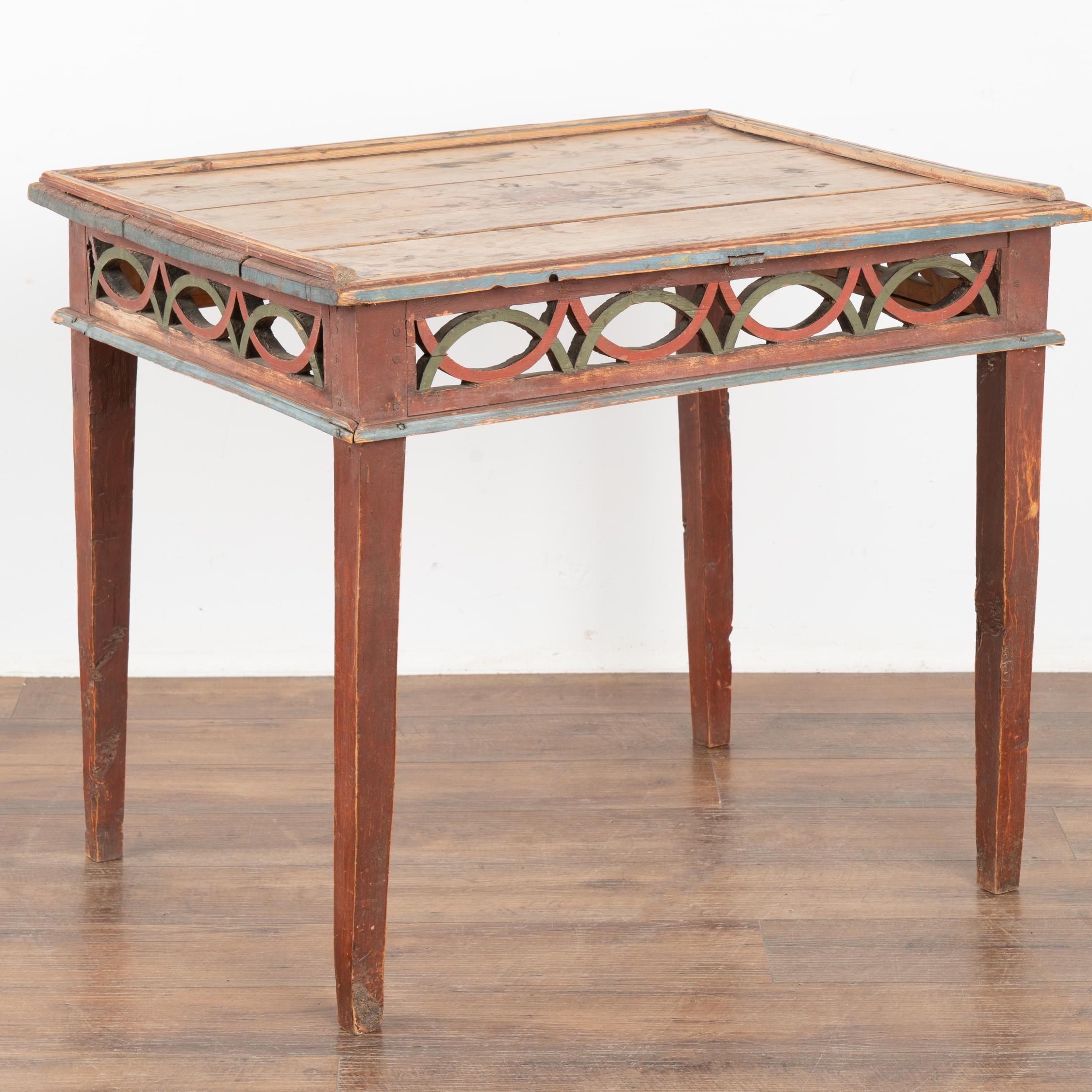 The paint is all original on this pine side table with tapered legs from Sweden. The decorative carved/lattice skirt (along all 4 sides) in red and green compliment the brick red base with blue trim.
While the top is natural pine, the residual