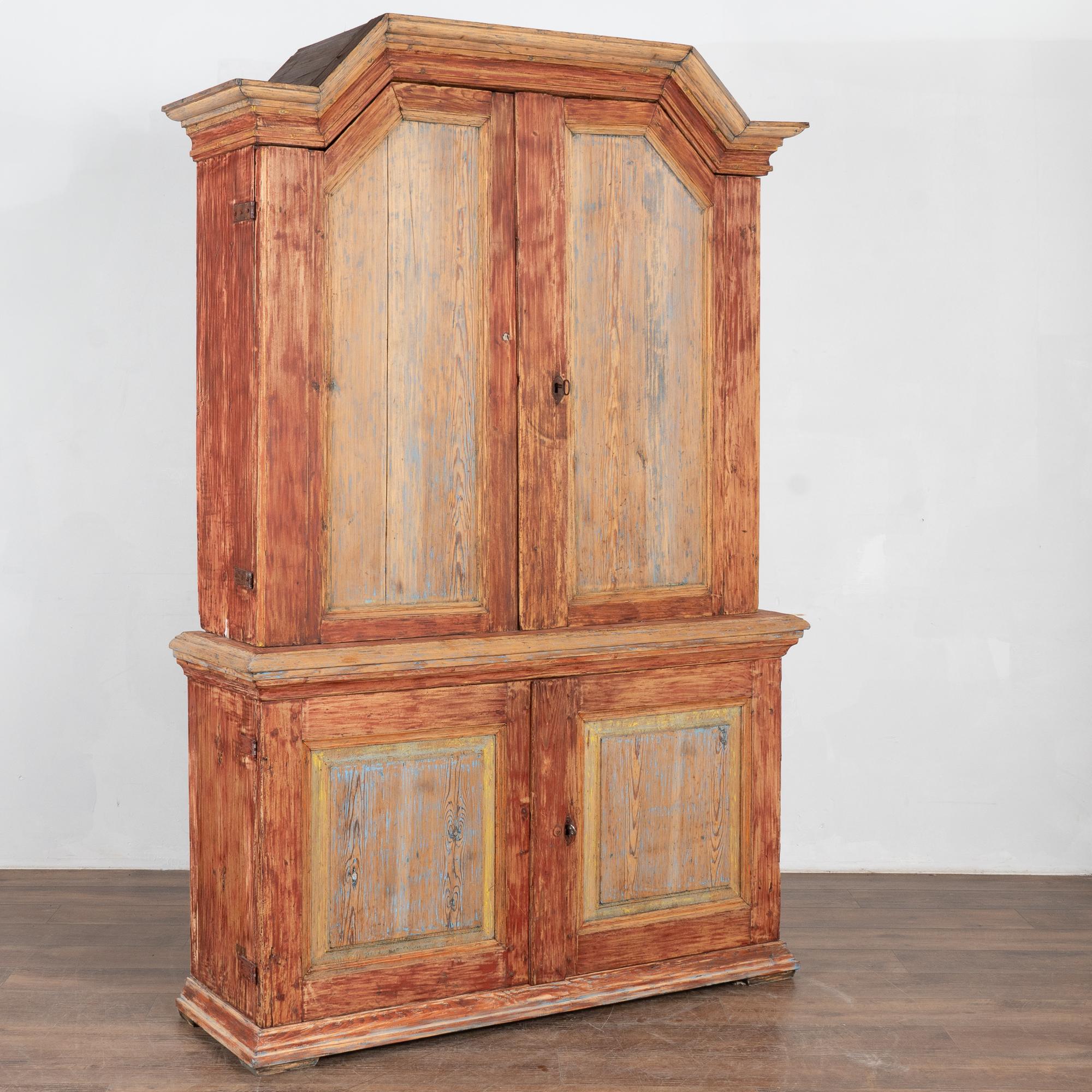 Cette armoire en pin présente la patine authentique que beaucoup recherchent dans les meubles du début des années 1800. Le fond original peint en rouge bruni est complété par des panneaux bleus bordés de jaune. La finition a été grattée, ce qui