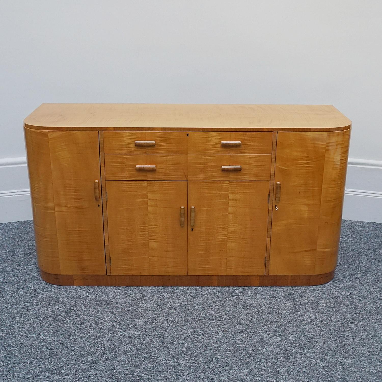 A curved Art Deco sideboard. Satinwood veneered with figured walnut banding and original walnut handles. Two central upper drawers with felt lined cutlery top drawer. Lower central cabinet opens to reveal deep shelved section with two shelved