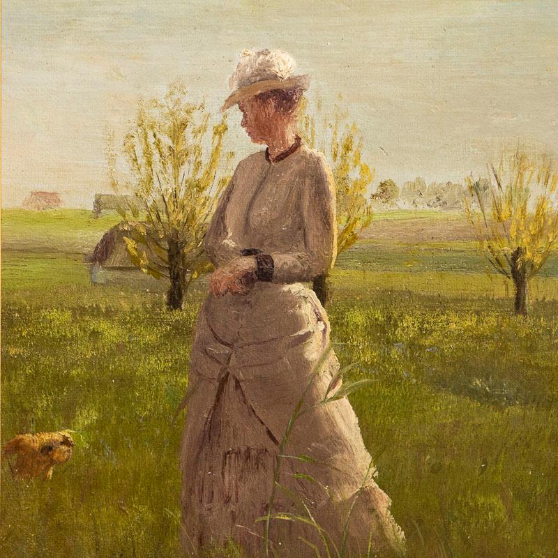 Cette belle peinture à l'huile originale représente une jeune femme élégante lors d'une journée d'été à la campagne. Regardez bien, elle semble avoir un petit chien à ses côtés. Bien que le peintre soit inconnu, cette petite peinture a été