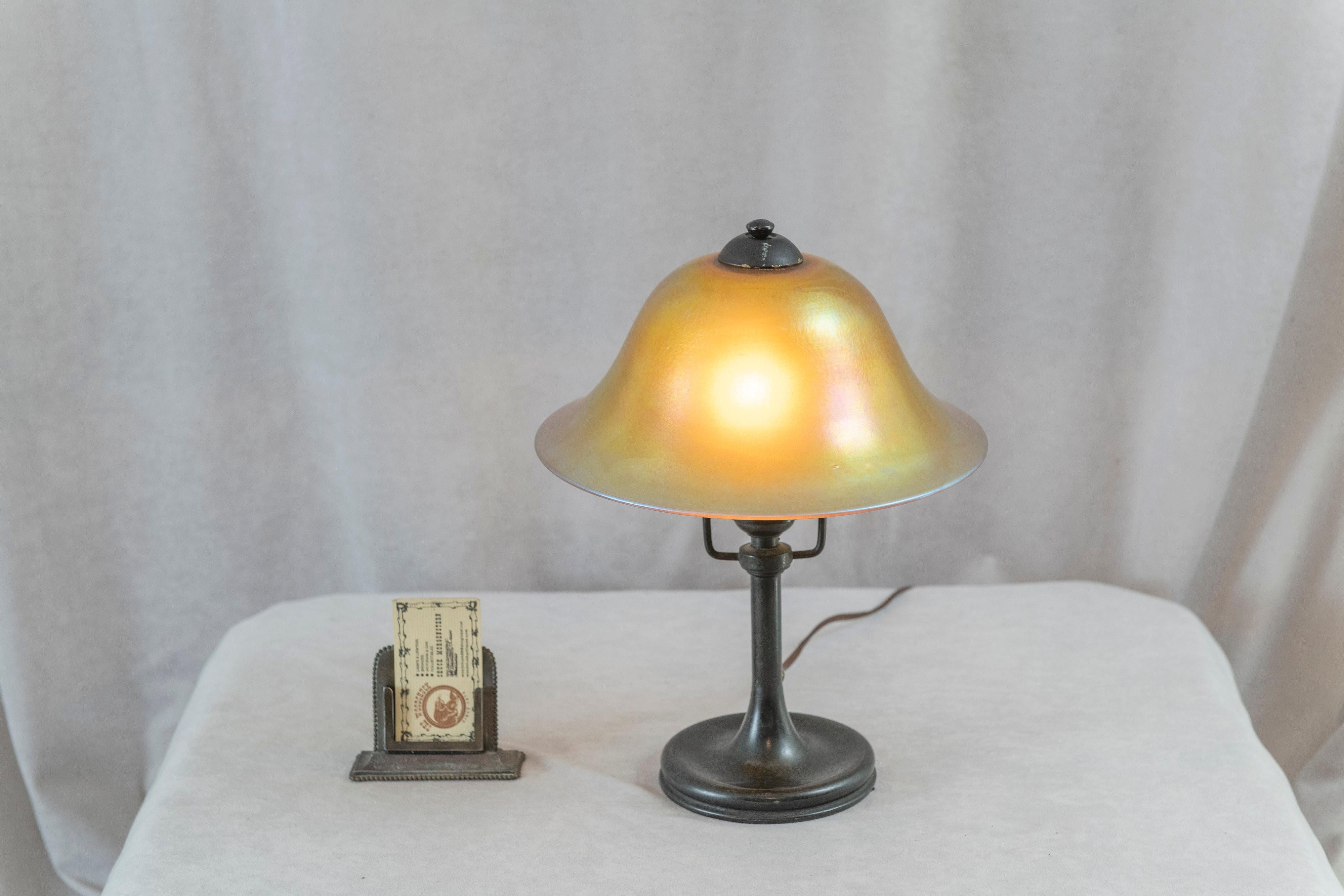  This sweet little lamp was made by one of the premier glass makers, Steuben. The shade is hand blown and displays an iridescent quality. The base is richly patinated bronze. We must emphasize that this is a completely original package. The shade