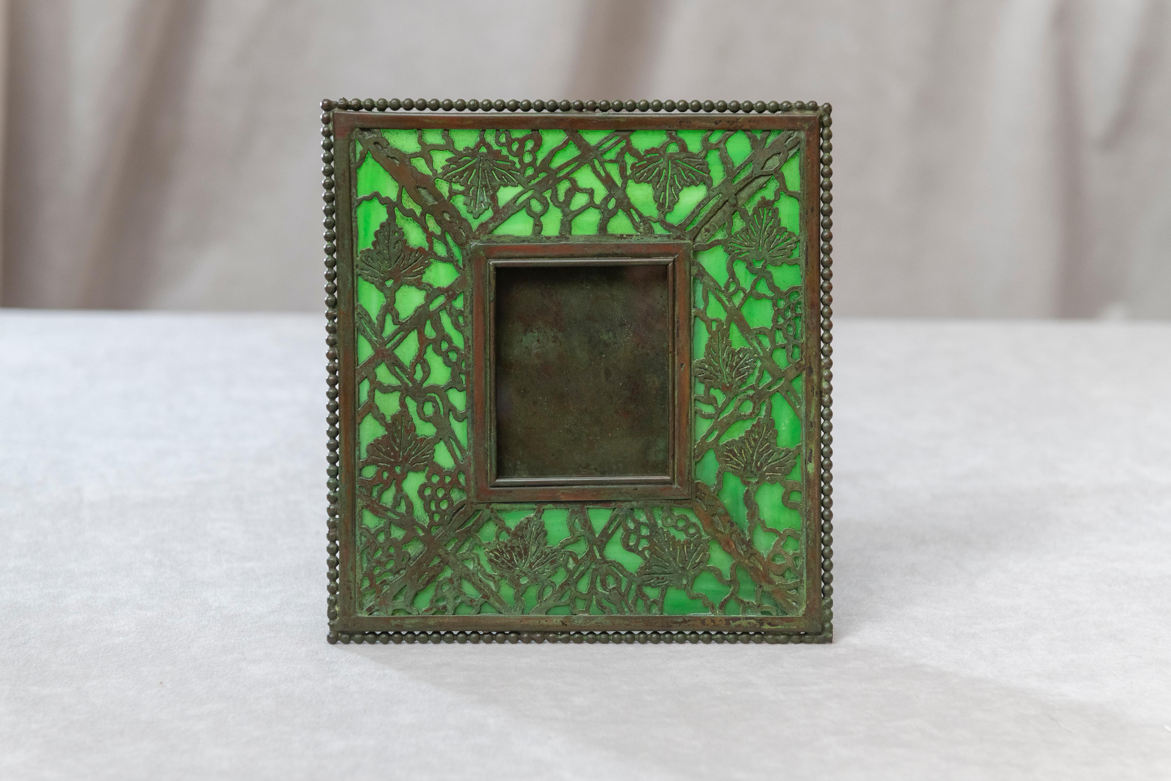 This smaller example of Tiffany's typical picture frame is a fine example of the genius of the man. Done in the grapevine pattern with green glass and finished in a rich warm patination to the metal. A lovely choice frame that can be fitted in most