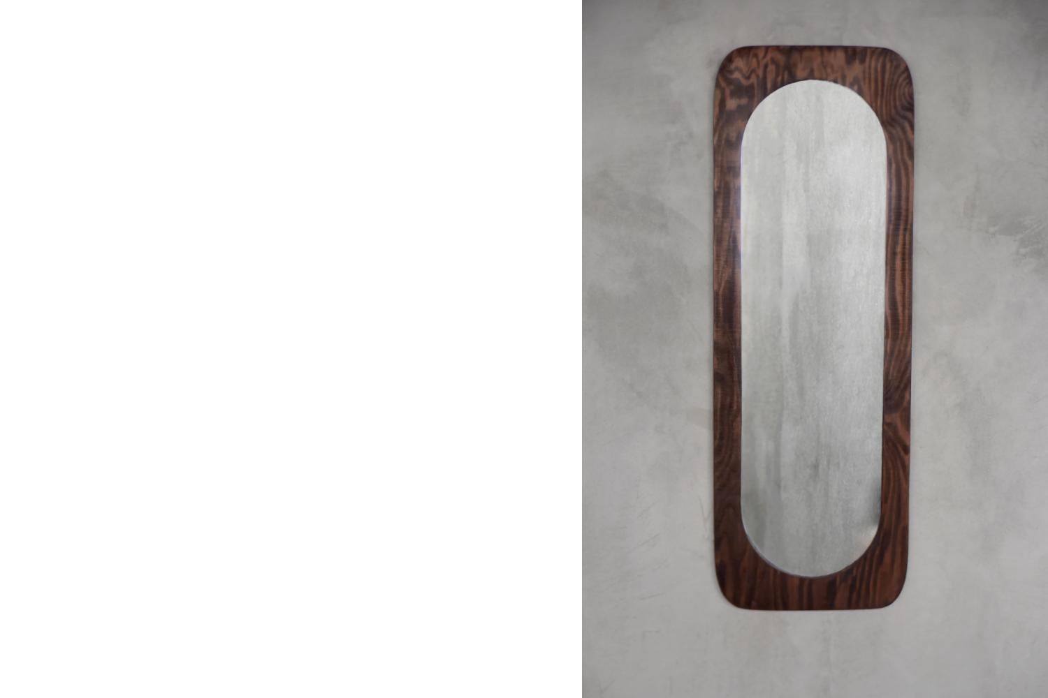 This tall wall mirror was made in Denmark during the 1950s. The rectangular mirror frame with flowing edges is made of solid wood in a dark shade of brown and tapers downwards. The slender form of the mirror makes the object light, which reflects