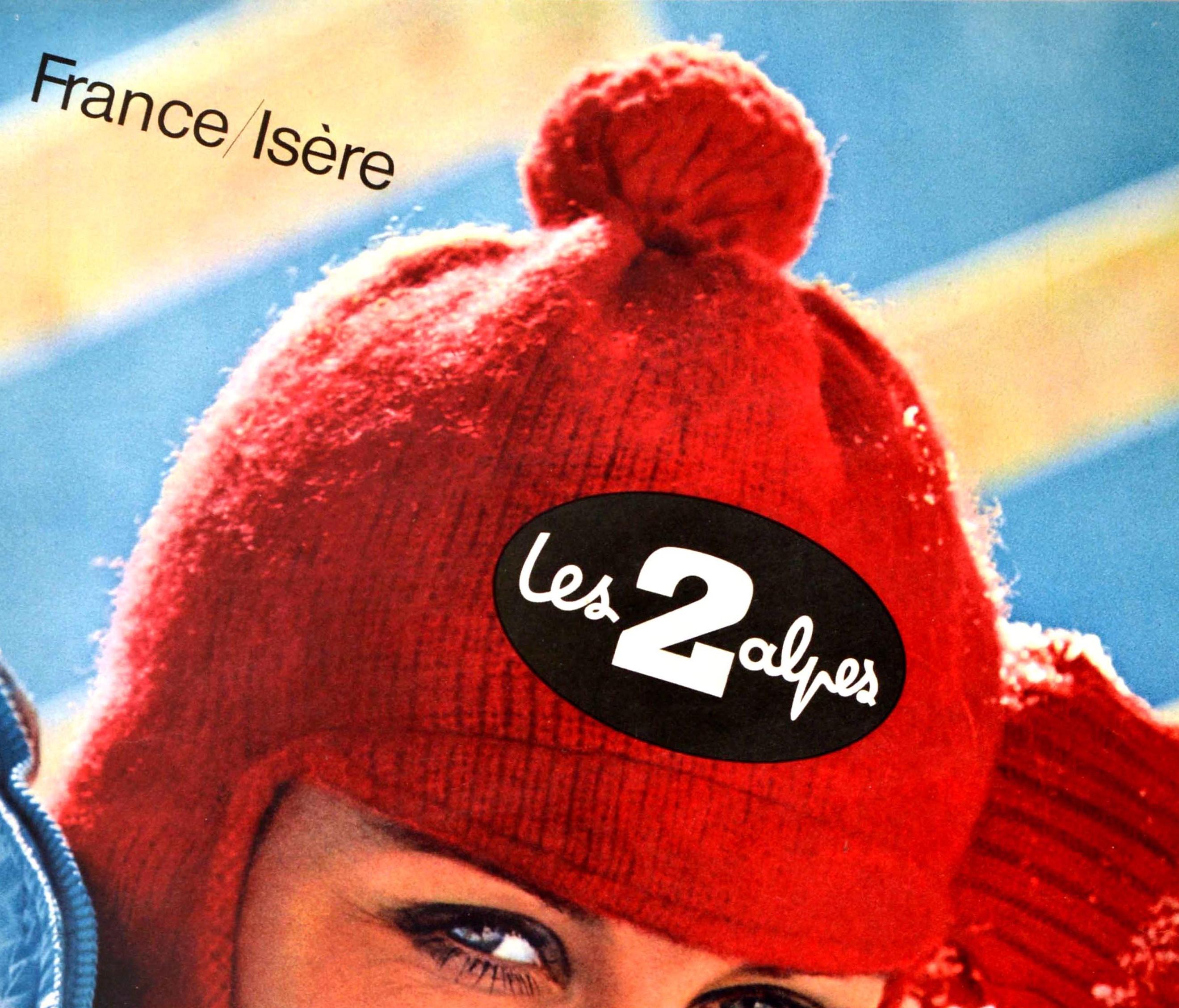 Original vintage winter sport travel poster for France Isere Les 2 Alpes ski resort featuring a great photograph of a lady wearing red wool balaclava hat and gloves and a blue ski jacket resting on the snow and smiling to the viewer. The popular
