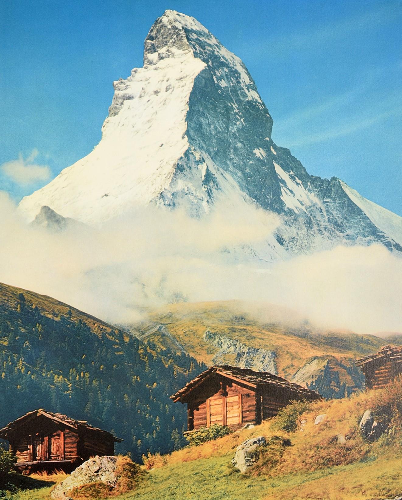 Affiche de voyage originale d'époque pour Zermatt:: en Suisse:: montrant une vue pittoresque de la montagne du Cervin:: coiffée de neige:: à travers une fine couche de nuages:: depuis des chalets en bois et des rochers dans un champ en contrebas.