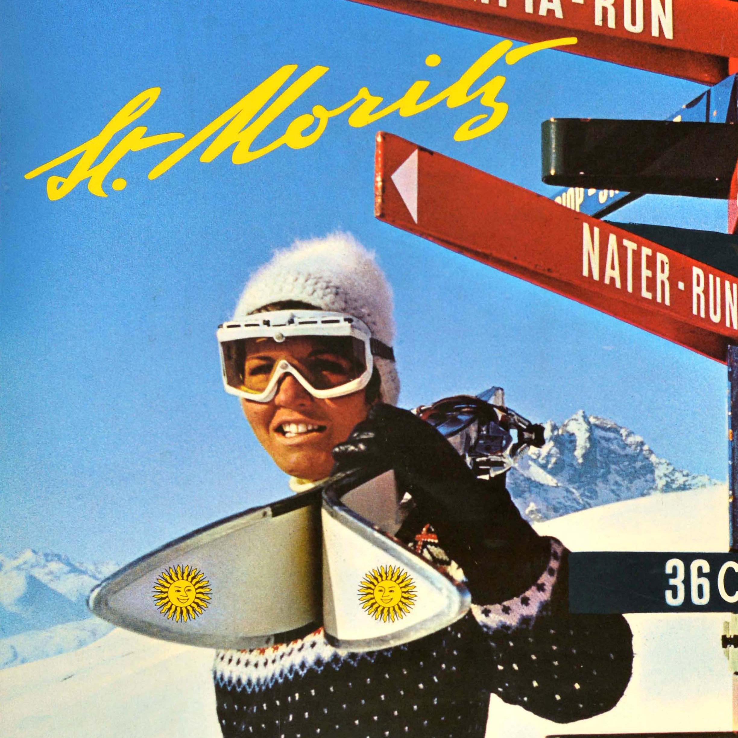 Original vintage skiing poster for the alpine resort of St Moritz in Switzerland featuring a skier wearing ski goggles, a bobble hat and knitted jumper, and holding poles and a pair of skis with the St Moritz sun logo on them, standing in front of a