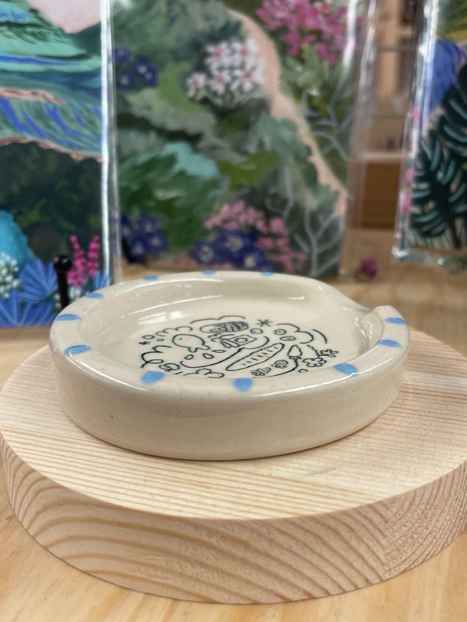 This spoon rest has an adorable cartoon doodle in the center. Makers mark on the bottom. Little Oakie Studios is a Seattle based company specializing in Kawaii inspired design. Every piece is Handmade, with Slight variation natural to the process of