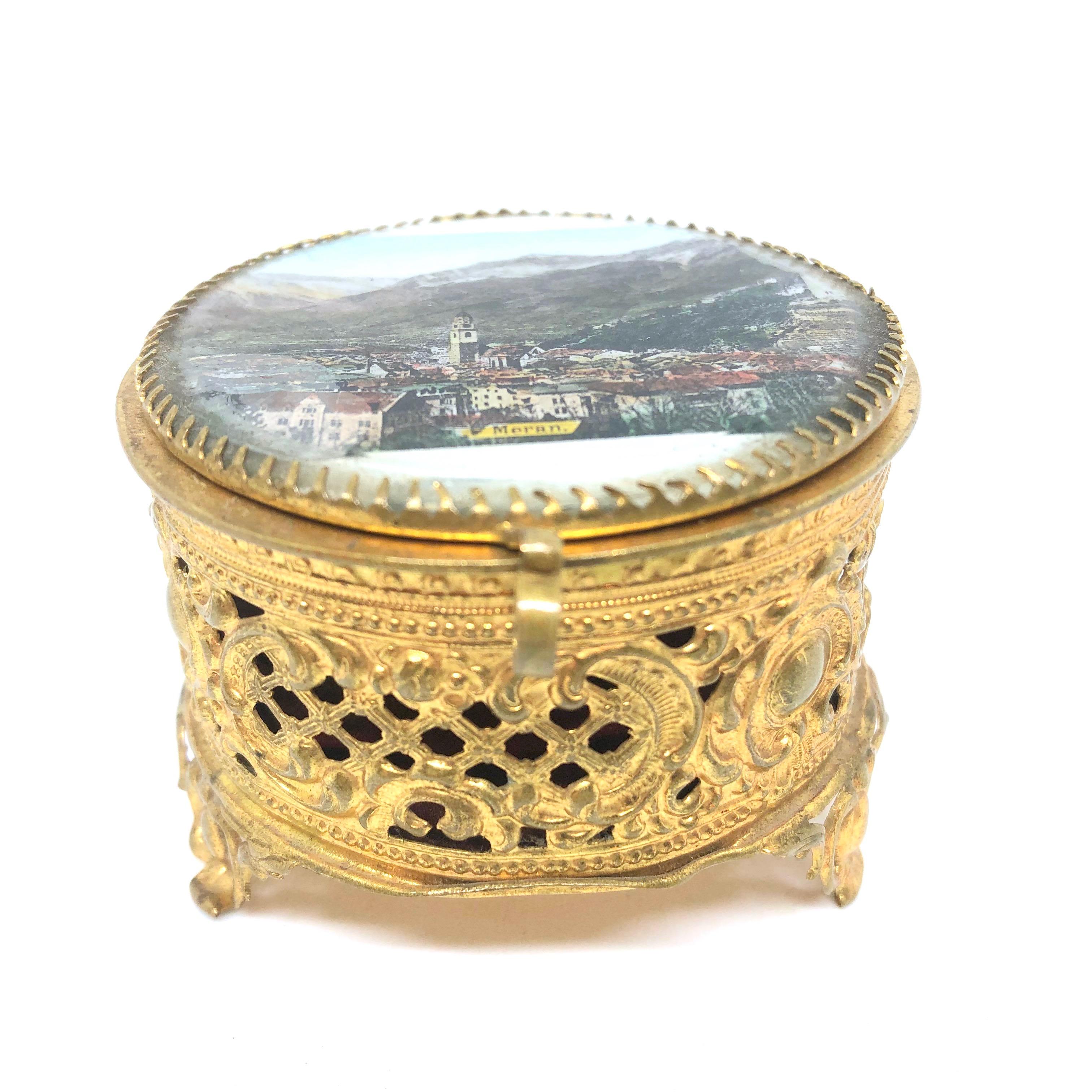 Una preciosa caja de baratijas con tapa de cristal hecha de ormolu. La parte superior abatible presenta una vista de la ciudad de Meran. No se ha llevado a cabo ninguna restauración en esta encantadora cajita de baratijas, que permanece en