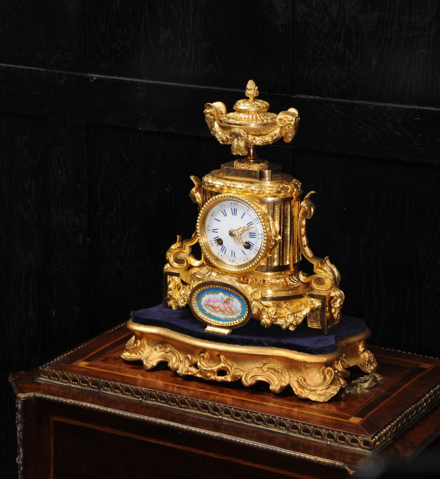 A lovely ormolu boudoir clock, antique French dating from circa 1860. It features a charming porcelain panel, delicately painted with a cherub in the manner of Sèvres. It is classical in style with scrolls and a large urn with rams head handles to