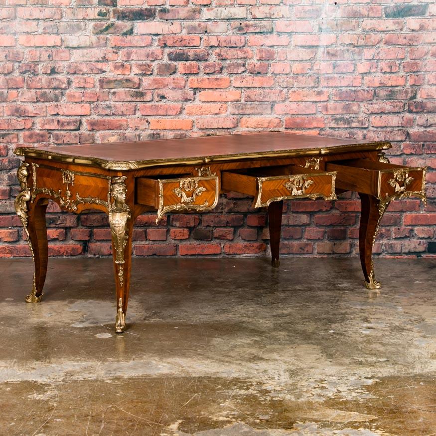 This remarkable 20th century French writing desk or 'bureau plat' in the Louis XV style, is fitted with ornate brass ormolu and a handsome hand tooled leather top. The desk is free standing which allows one to appreciate the intricate wood inlay and