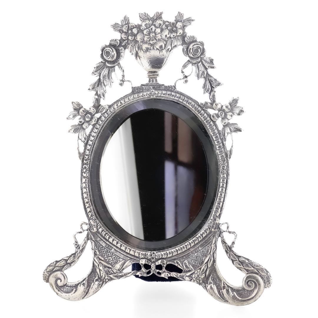 A fine vintage silver dresser or vanity mirror.

In .830 solid silver.

In an oval form with beveled silvered glass. Supported by two bracket feet and surmounted by a vase with a floral bouquet.

Having an easel back covered in blue velvet.

Marked