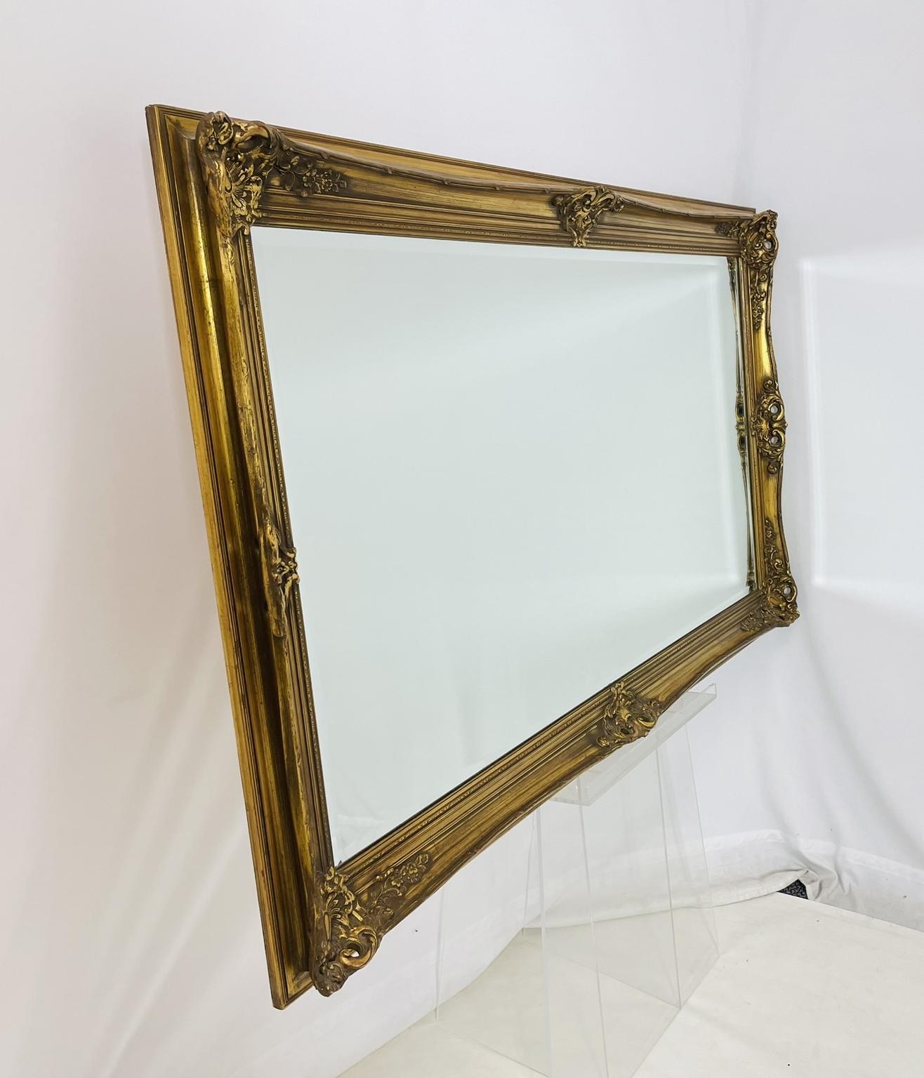 Un grandioso y ornamentado espejo de pared rectangular fabricado en Bélgica en la década de 1960, con un elegante marco de madera dorada ricamente adornado con tallas florales y volutas de estilo rococó. Los detalles escultóricos están ejecutados en