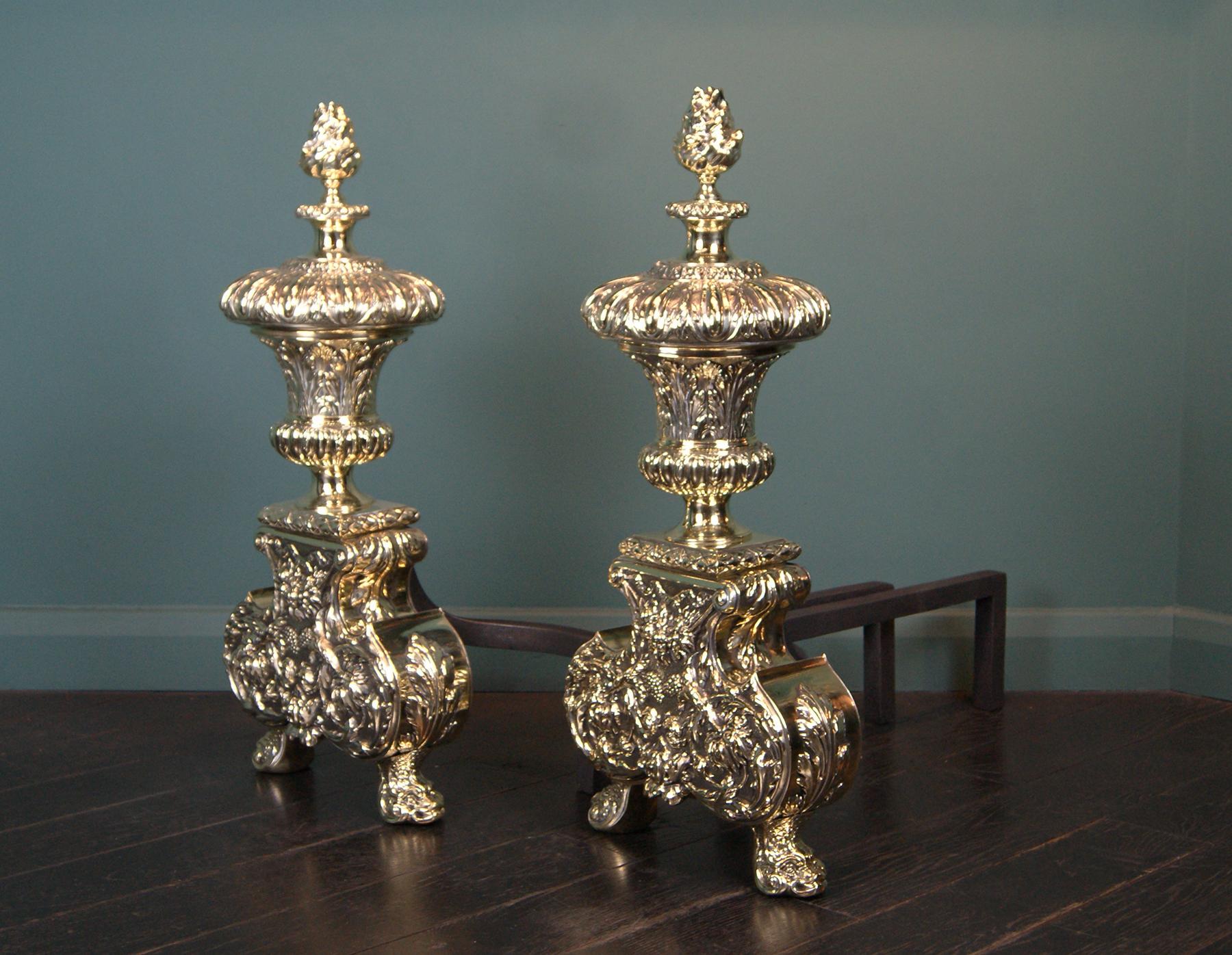 A beautifully-made pair of brass English andirons, with gadrooned urns surmounted by flame-topped finials. The substantial base supports with scrolled foliage and fruit in relief, on stylised dolphin feet. This set of andirons are near identical to