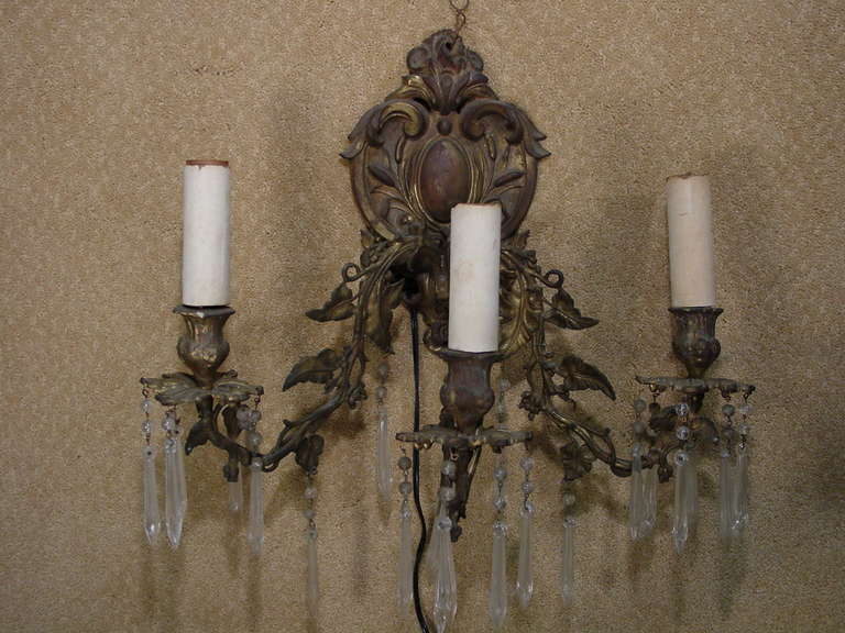 This bronze Rococo wall sconce features three arms. Each of the three arms are entwined with bronze vines and leaves that extend from the ornate wall mount. The light fixtures themselves feature prismed crystal pendants.
