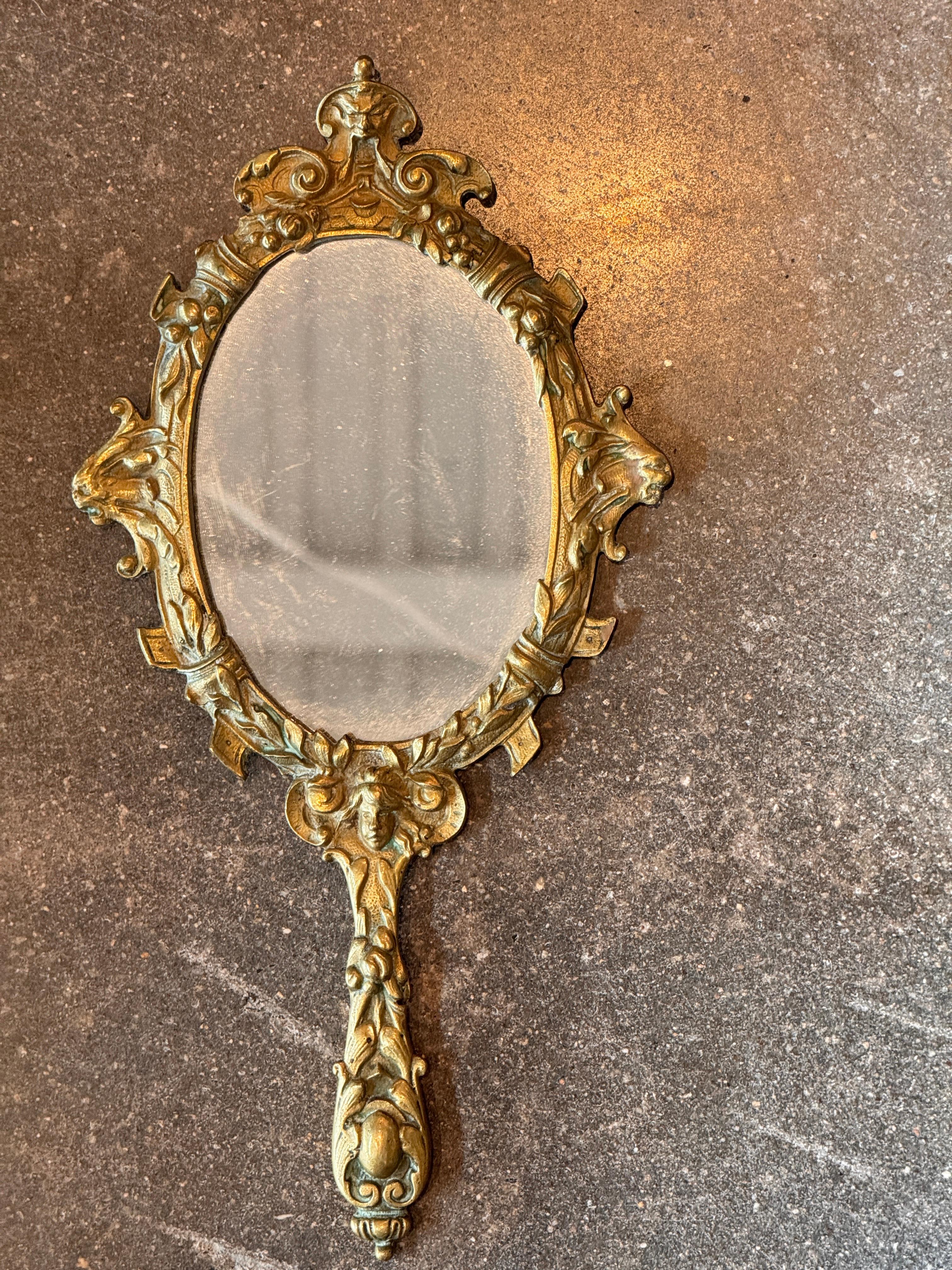 Miroir à main en laiton coulé de manière élaborée, présentant des motifs figuratifs et feuillagés, dont un visage central classique à la base et des rinceaux dans tout le cadre. La surface présente une usure et une patine adaptées à l'âge qui