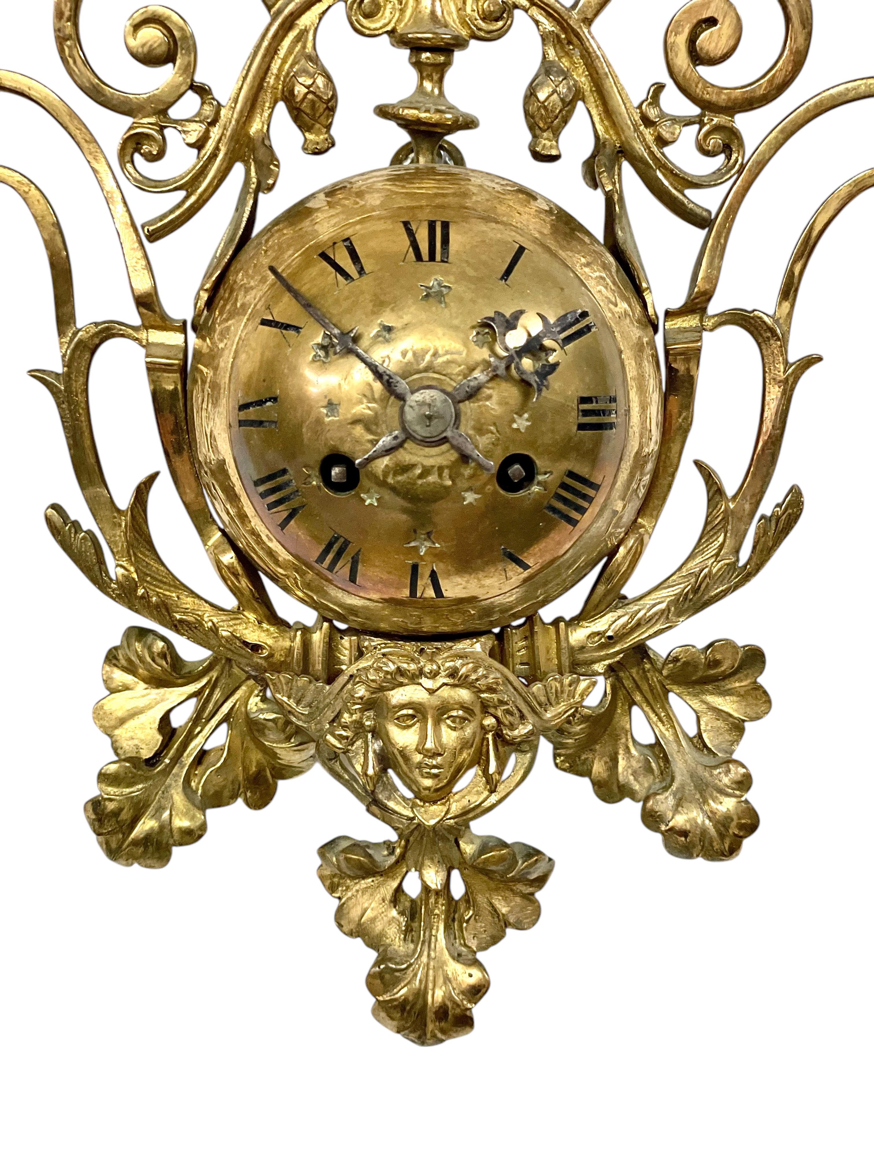 Diese charmante Wanduhr aus vergoldeter Bronze aus der Zeit um 1900 ist ein exquisites Beispiel für dekorative Handwerkskunst aus dem späten 19. bis frühen 20. Jahrhundert. Sie ist sowohl ein funktionaler Zeitmesser als auch ein dekorativer Akzent
