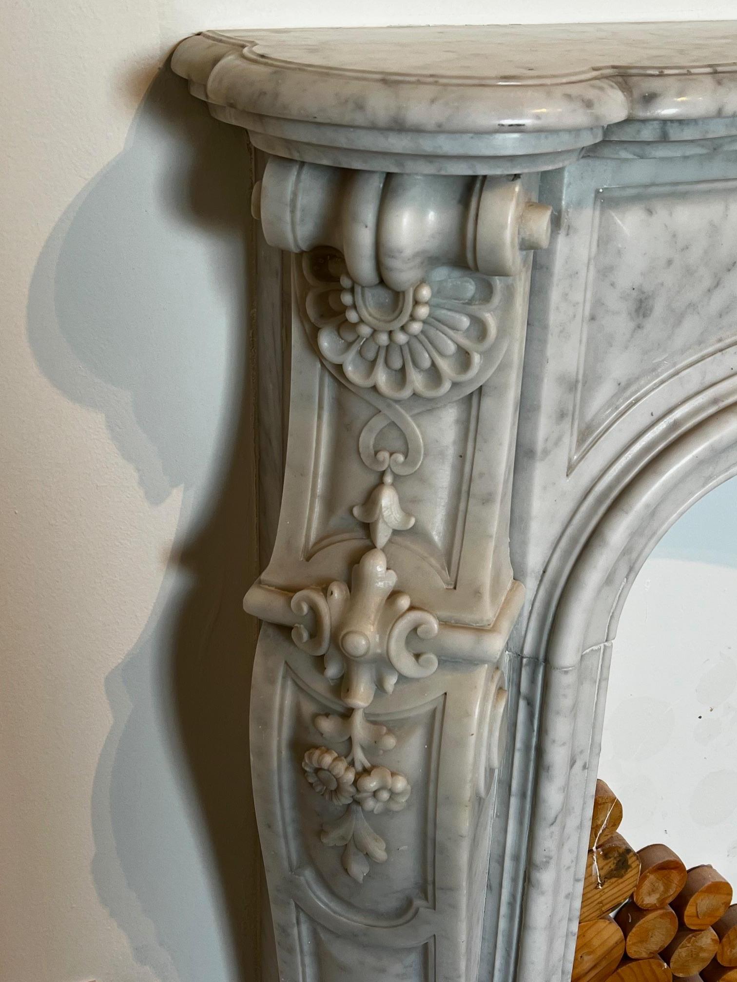 Dieser wunderschön verzierte Kamin, handgeschnitzt im Stil von Louis XV, ist aus halbpoliertem weißem Carrara-Marmor gefertigt. Sowohl auf dem Türsturz als auch auf den Beinen befinden sich fein geschnitzte florale Details. Handgeschnitzte Muscheln