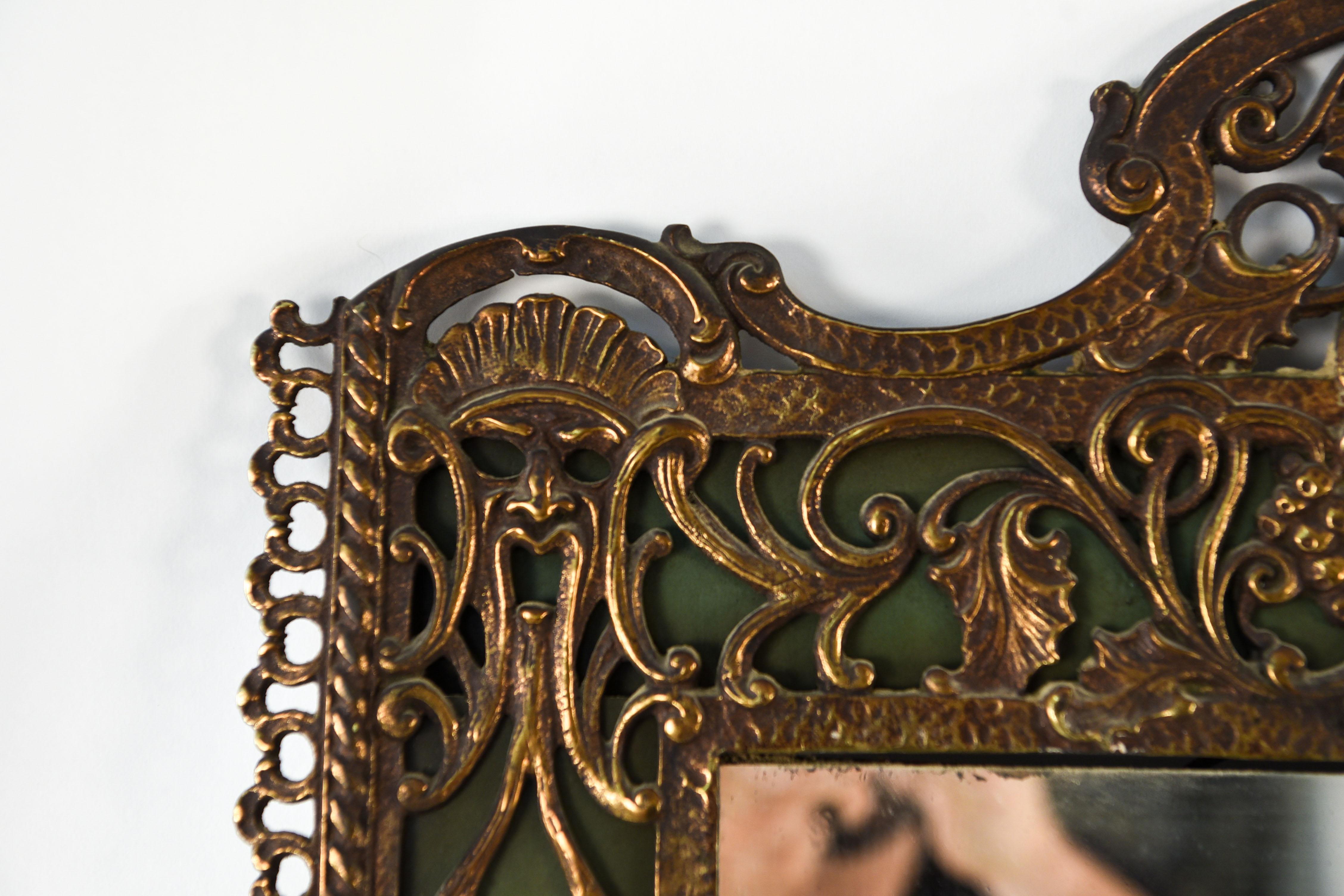 This ornate antique wall mirror features a frame with classical patterns including grape bunches and vines. Brass overlay on a green background.