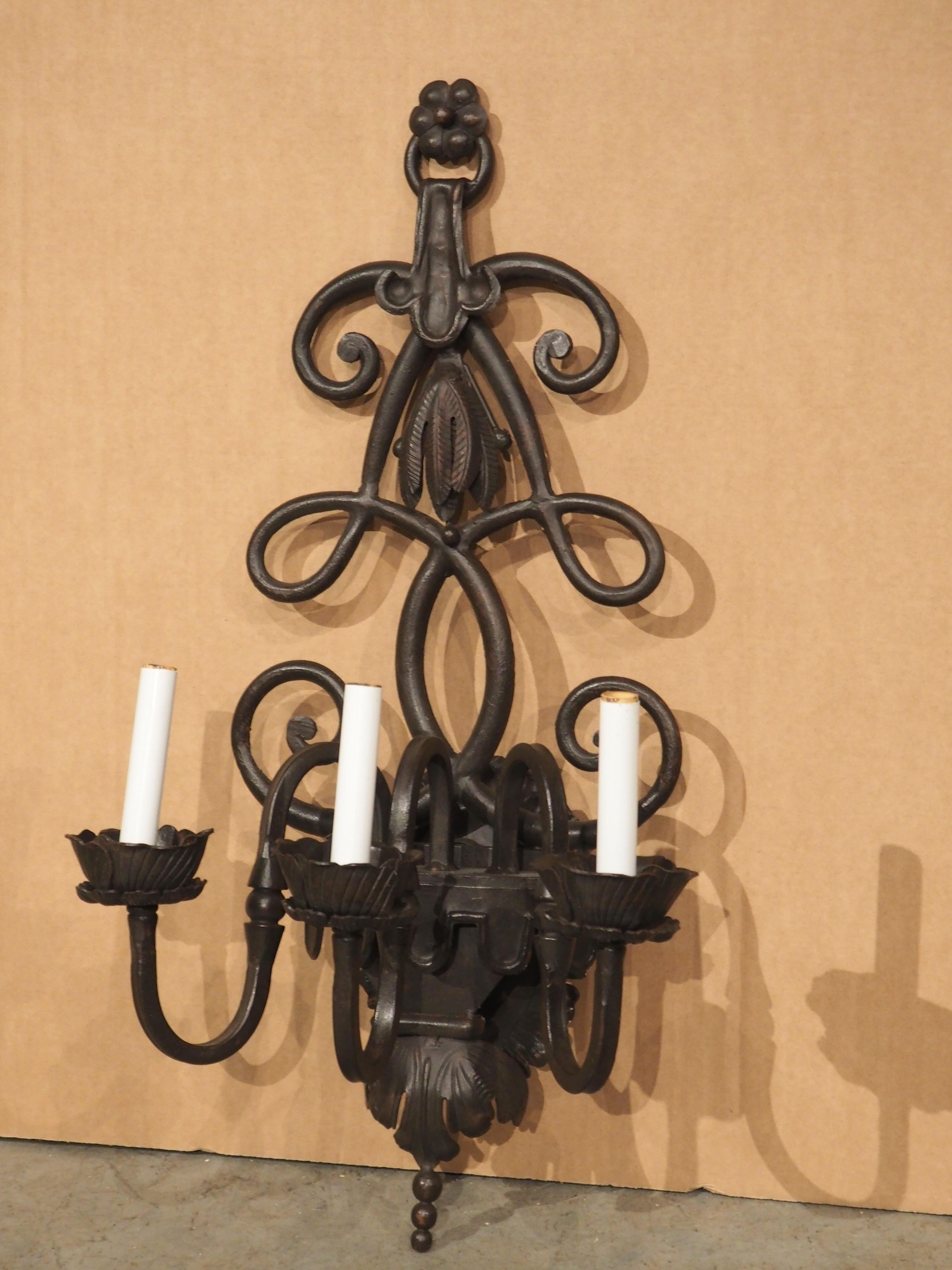 Dieser dreiarmige Wandleuchter aus Eisen ist im Stil spanischer Architekturbeleuchtung geschmiedet und wird von weißen, künstlichen Kerzenhülsen gekrönt, die aus Untertassen herausragen. Jeder Arm hat die Form einer gewundenen S-Rolle mit einem