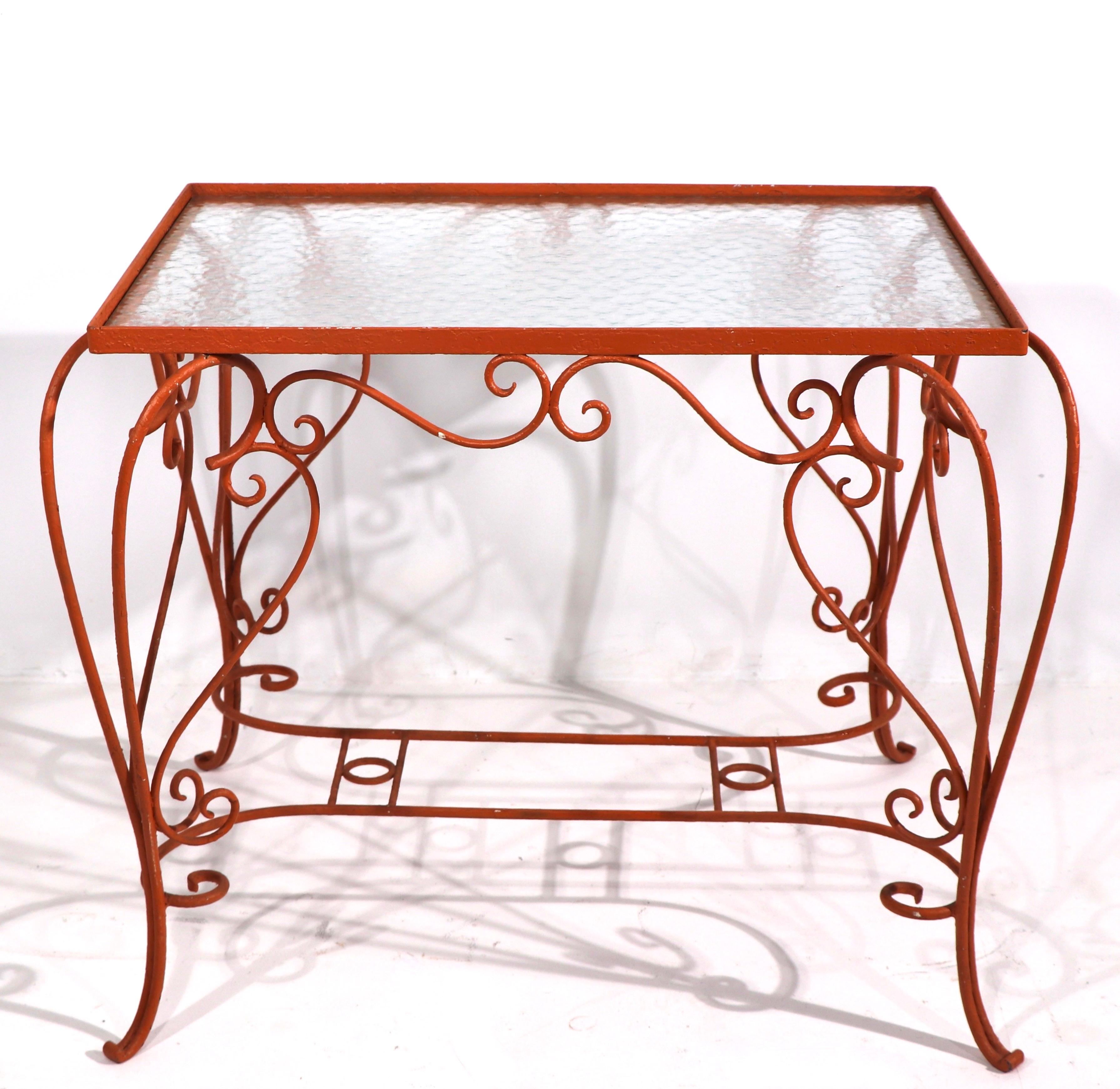 Charming wrought iron garden end, side table with ornate curlicue scroll work base and textured glass top. The table is currently in later orange/peach paint finish, usable as is or we offer custom powder coating if you prefer a different color, and