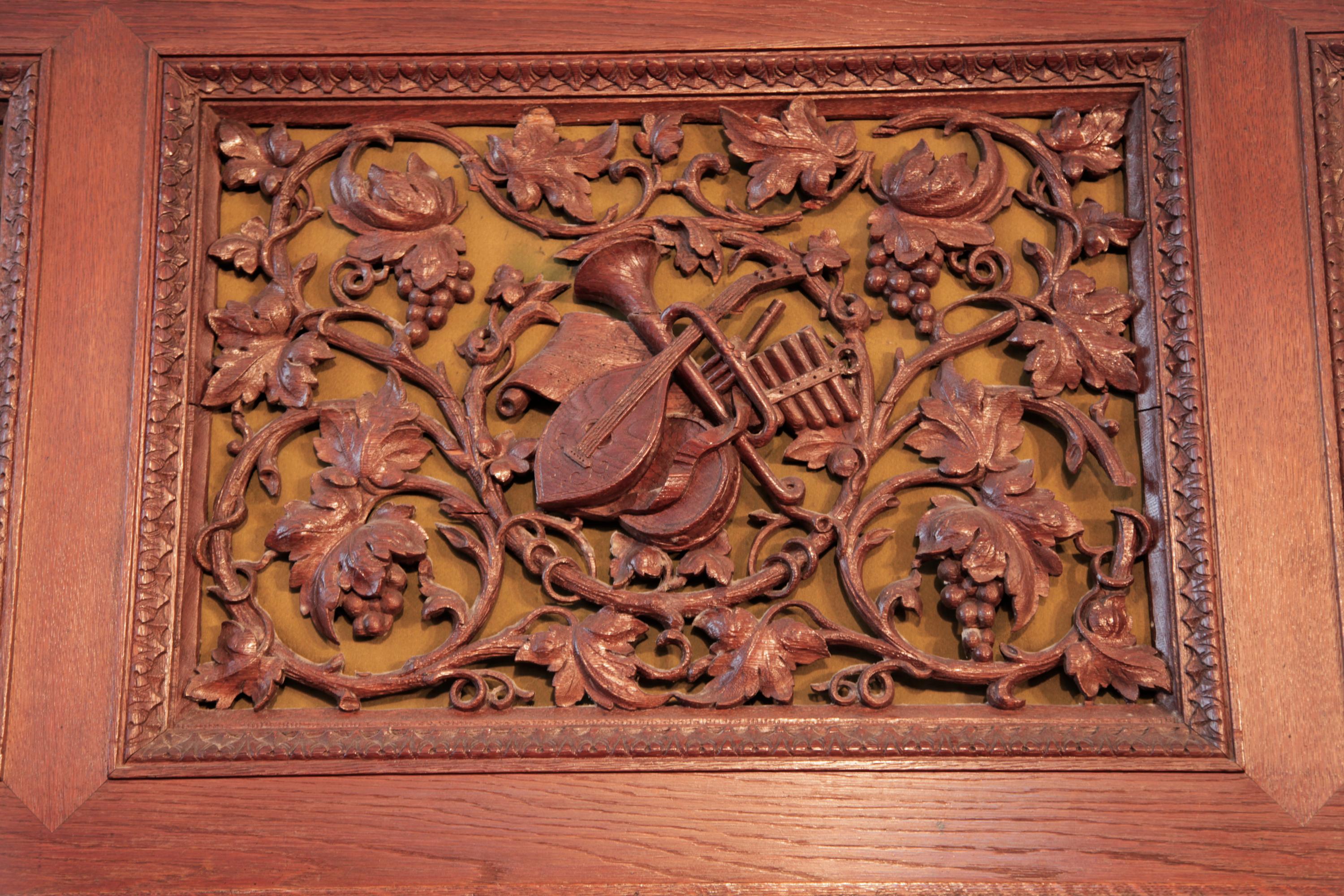 Piano droit Biese Hof avec caisse en chêne. L'ensemble du meuble présente des panneaux ajourés et sculptés en haut-relief, soutenus par un tissu doré. La face avant présente un ensemble d'instruments de musique entourés de vignes, de feuilles et de