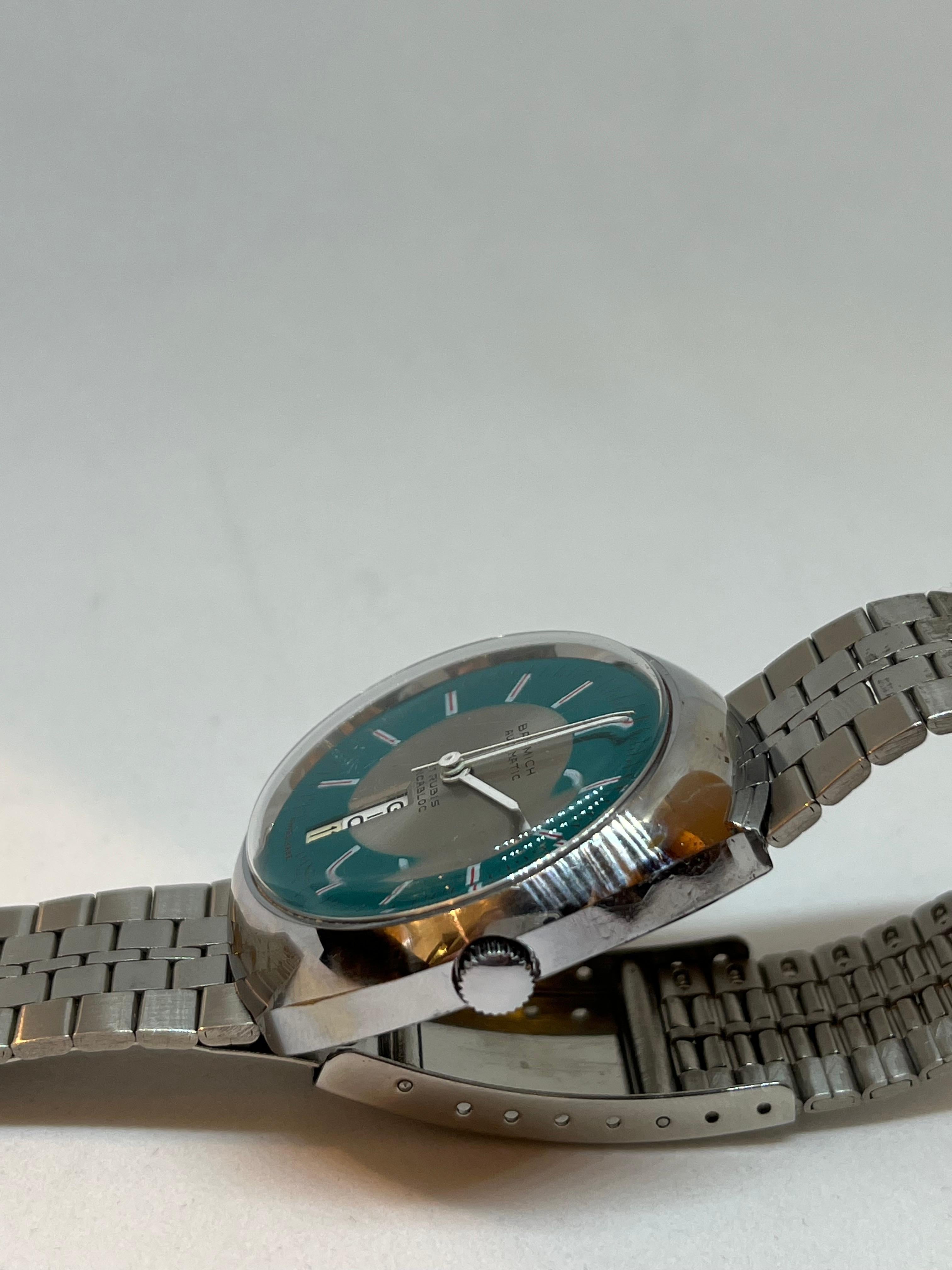 Este reloj es un elegante modelo automático vintage de la conocida marca Basmich.
Su diseño es típico de los años setenta y se aprecia en sus líneas suaves y redondeadas y en su caja cromada y pulida. 

La esfera verde azulado se llama 