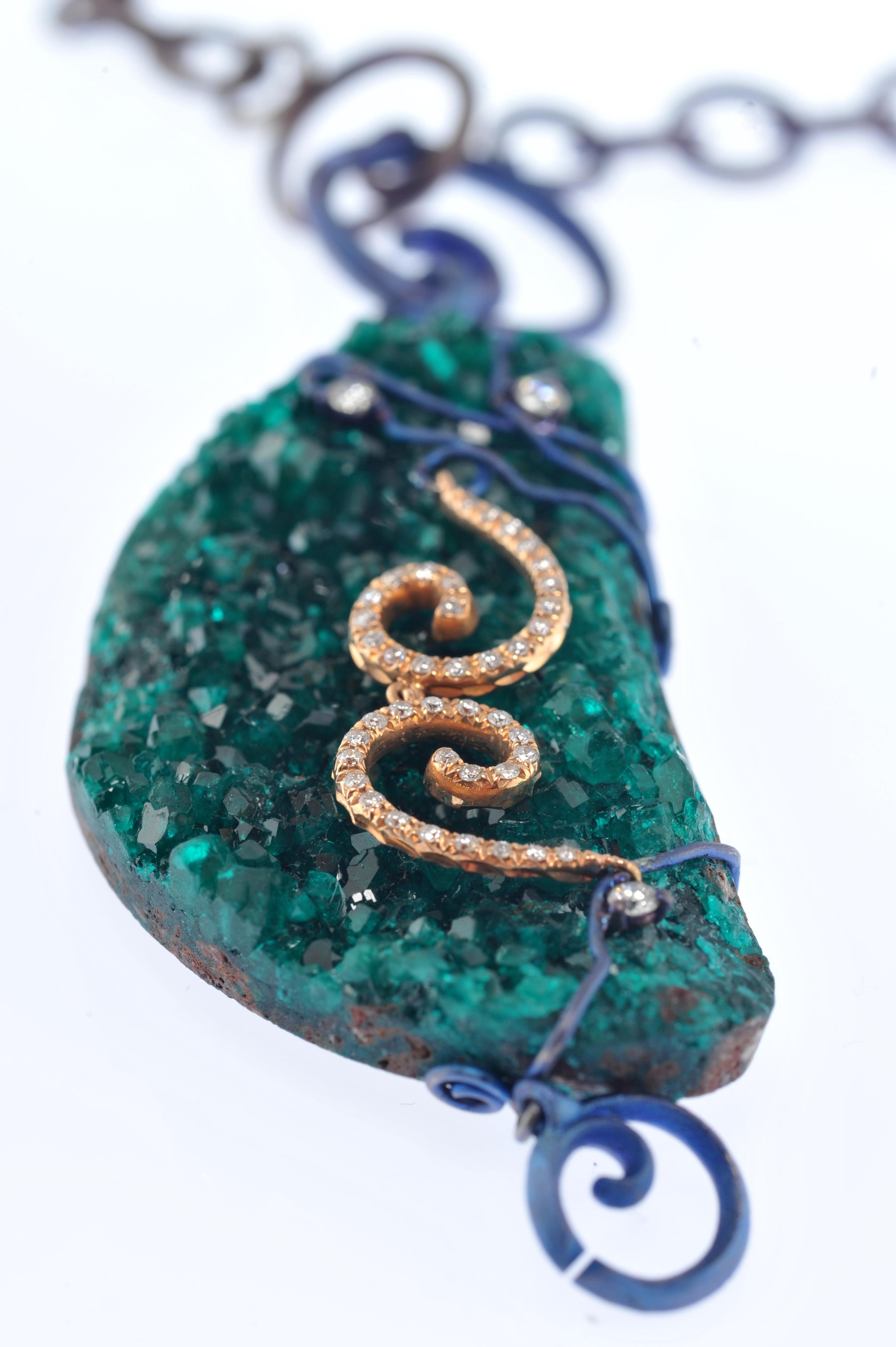 The pendant is realized with an uncut diopase, set with blue finish intertwined titanium wires. The stone is embellished with two 18Kt rose gold spiral and brilliant cut diamonds.
The pendant can be used on a rigid wire or with a chain as a