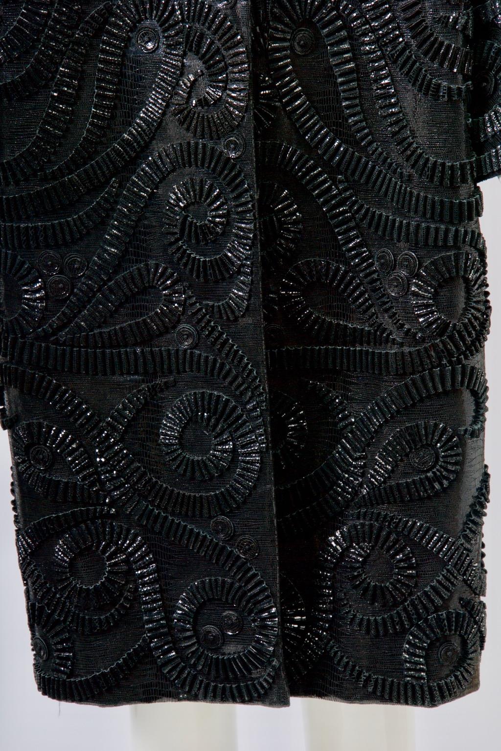 Schwarzer Lackmantel von Oscar de la Renta, vorne mit plissierten und gewirbelten Applikationen verziert, hinten undekoriert. Der Mantel hat einen einfachen, geraden Schnitt, ist kragenlos und hat handtellergroße Dolman-Ärmel. Eine strukturierte,