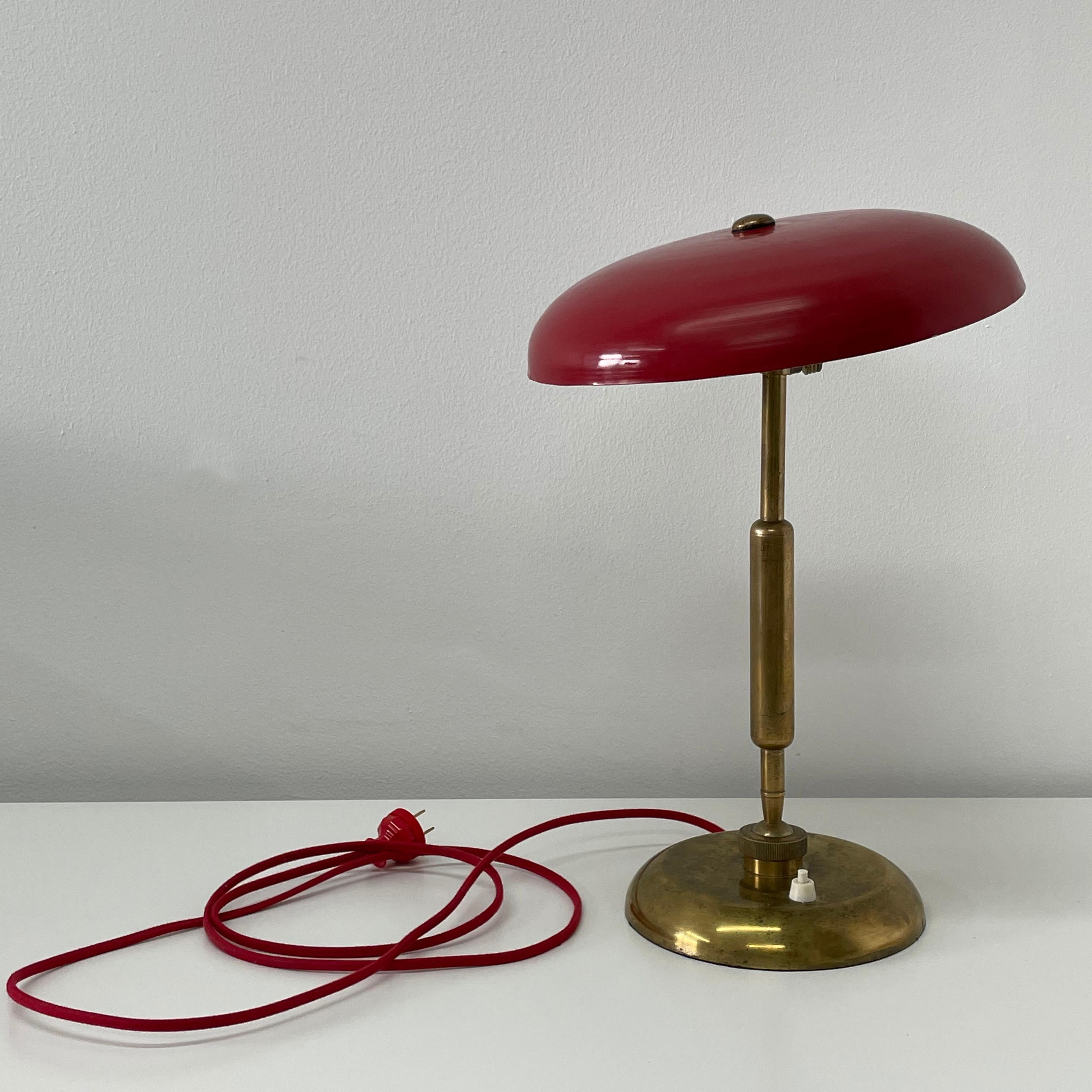 This brass lamp with lacquered cherry red shade designed by Oscar Tarlasco has a double ball joint that enables one to adjust the cant of both the stem and the shade. The lower joint has a handsome knurled collar detail. Its friendly shape,