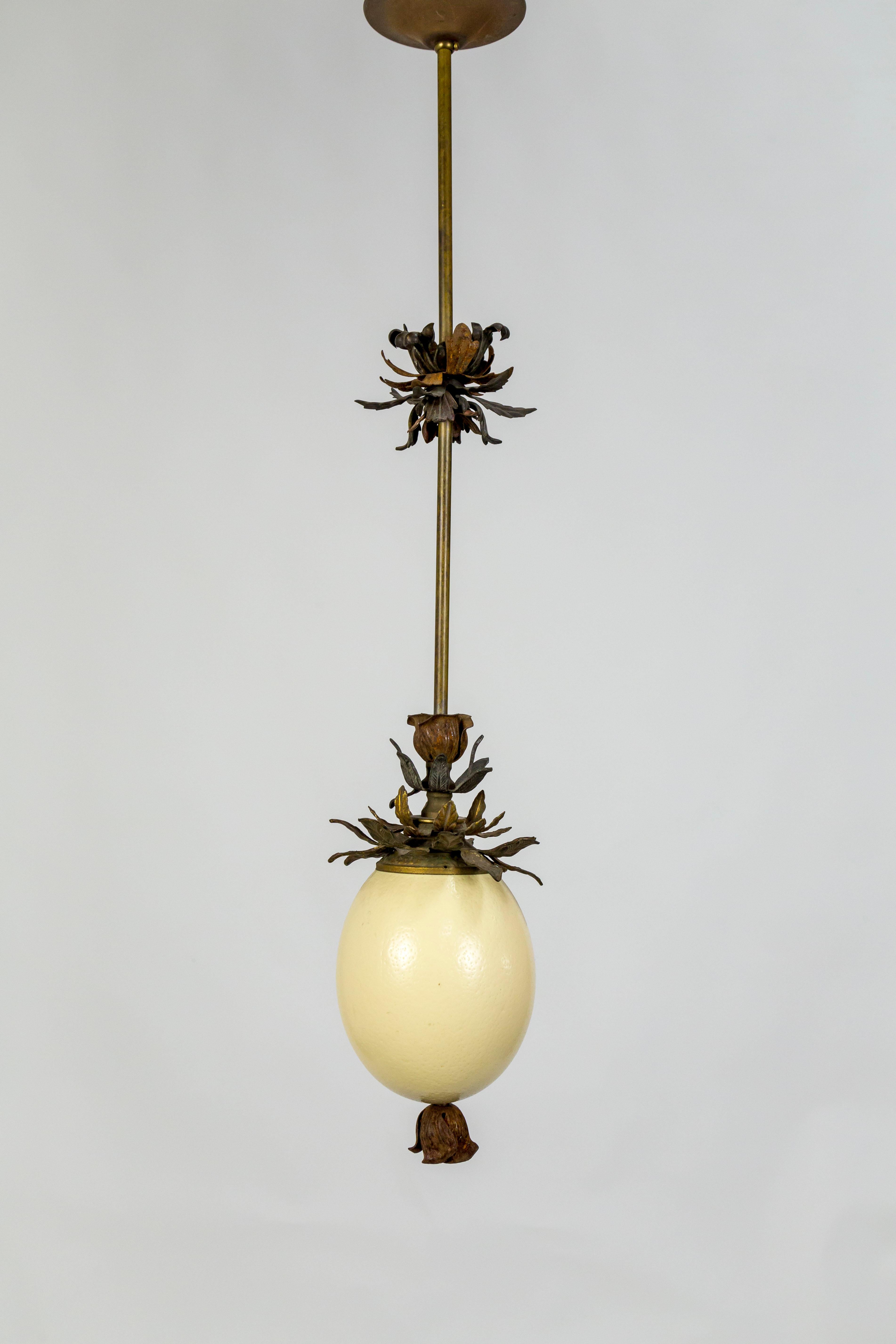 Gorgeous, handcrafted pendant light made with an ostrich egg and antique metal, floral decorative elements on a brass stem. The egg has an opaque-looking bone color that turns into a warm, varied tone when lit. Measures: 29.5